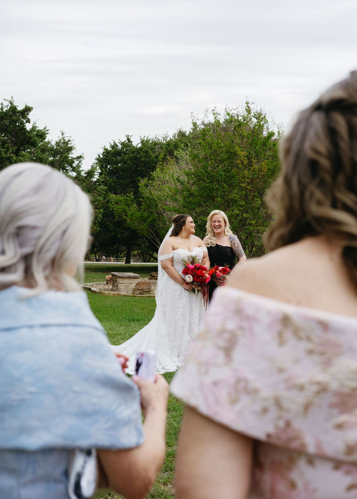 Two brides share a joyful moment outdoors at their wedding ceremony while guests watch and photograph them.