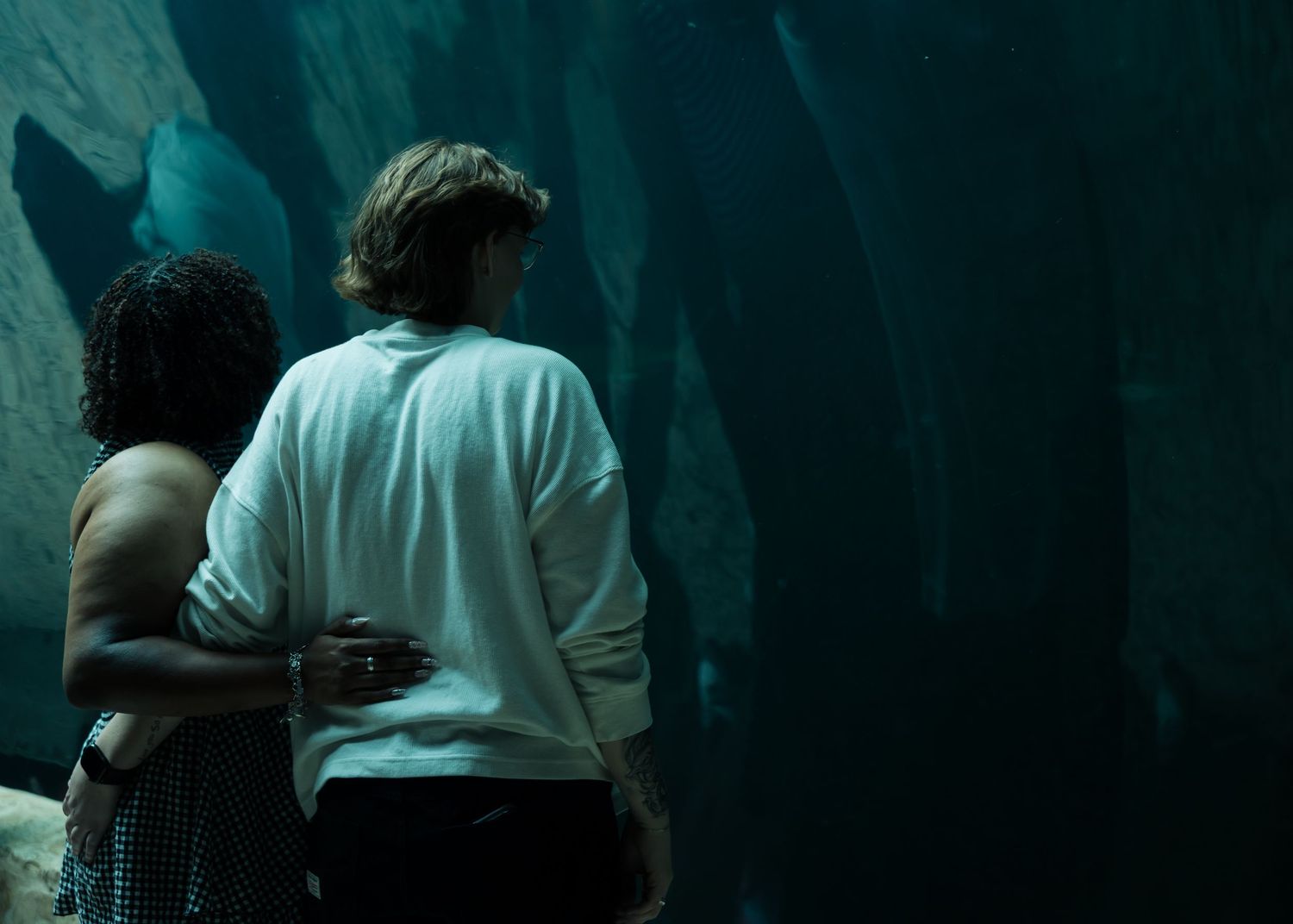 Couple embracing while viewing large aquarium tank with sharks in dim blue underwater lighting.