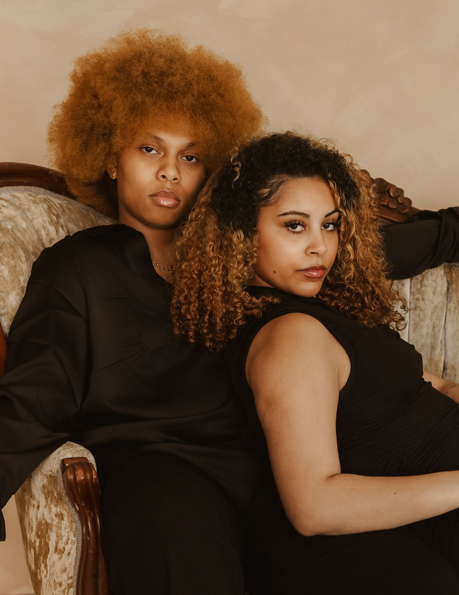 Two people with curly hair wearing black outfits pose together on a vintage sofa against a neutral backdrop.