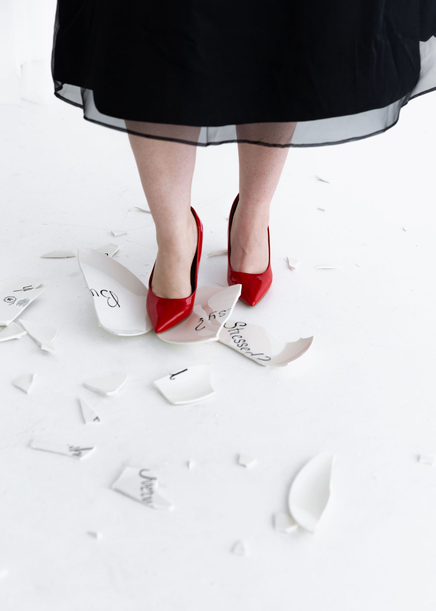 Red high heels and a black tulle skirt are photographed against a white floor scattered with paper confetti.