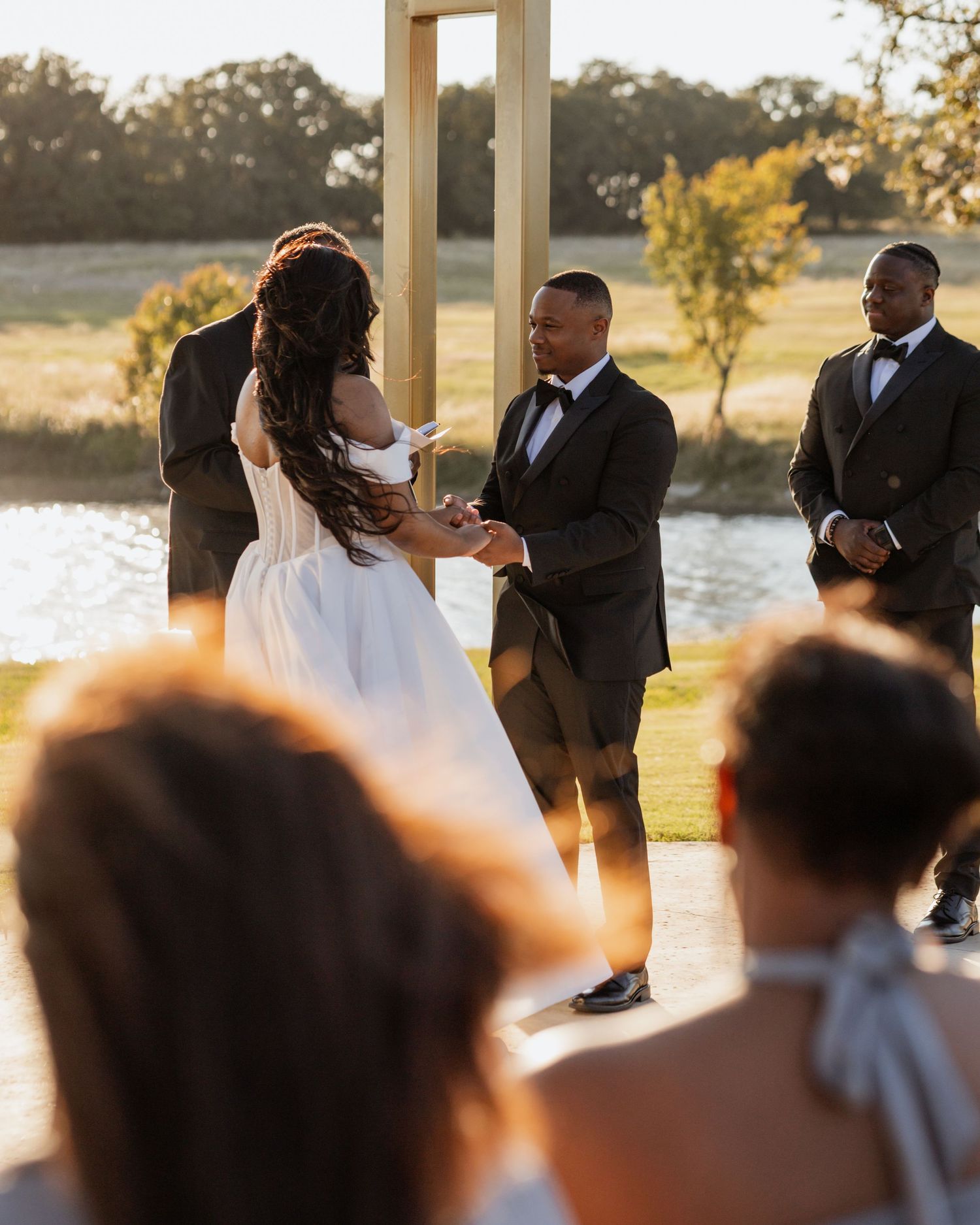 Wedding ceremony taking place at sunset by a lake with guests watching.