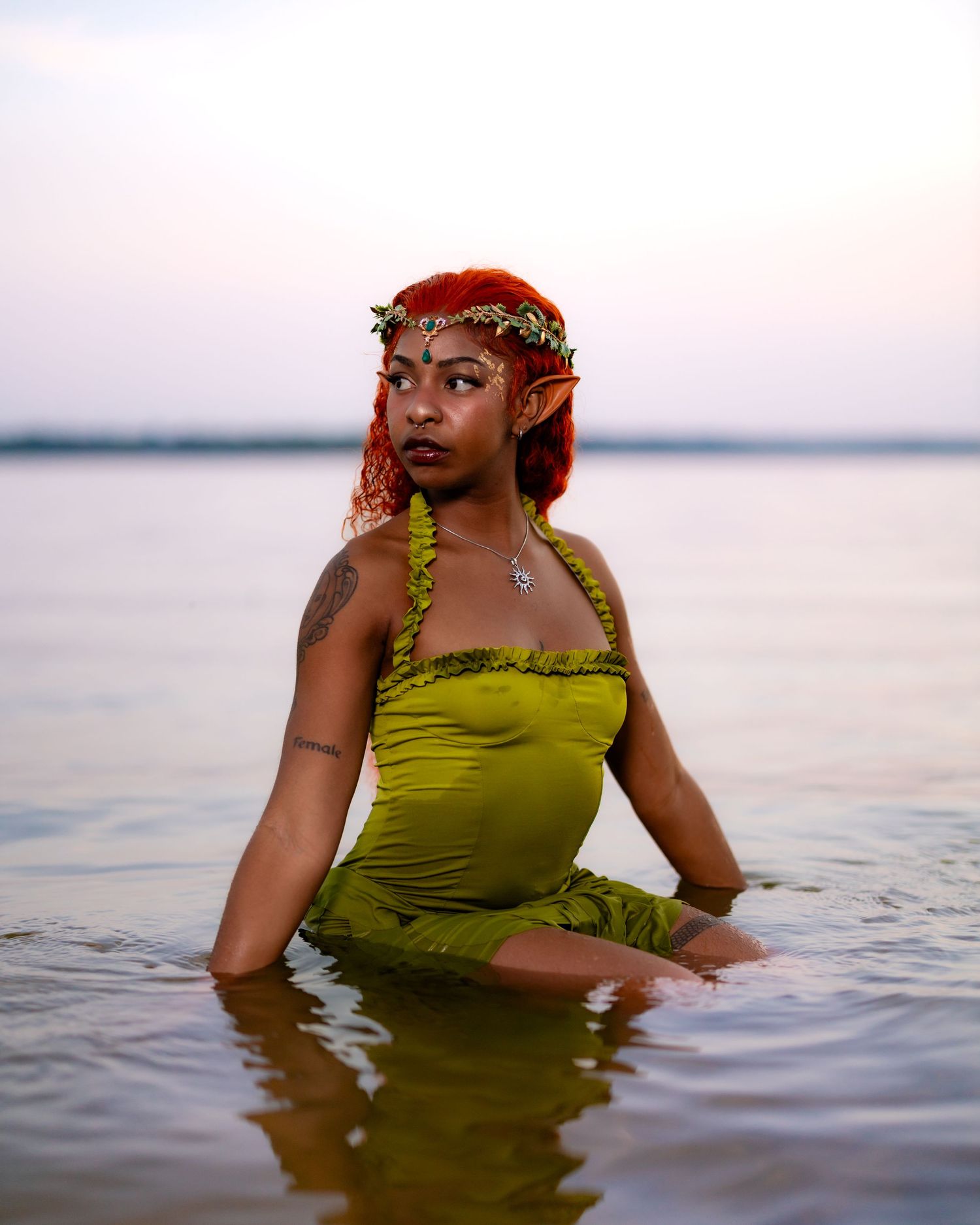 A mystical forest nymph in olive green dress and floral crown poses gracefully in tranquil waters at sunset.