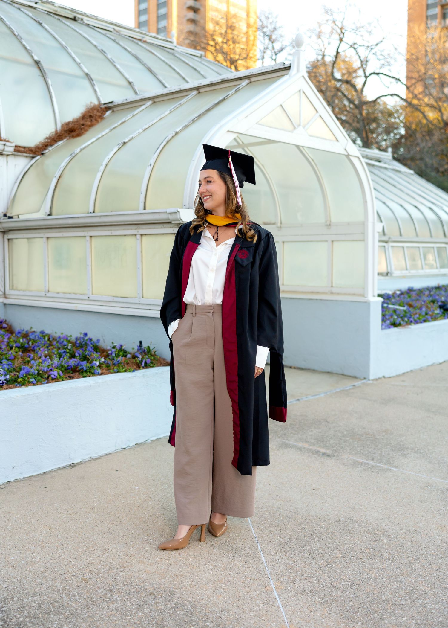 Graduate in academic regalia and tan pants posing in front of a greenhouse structure.