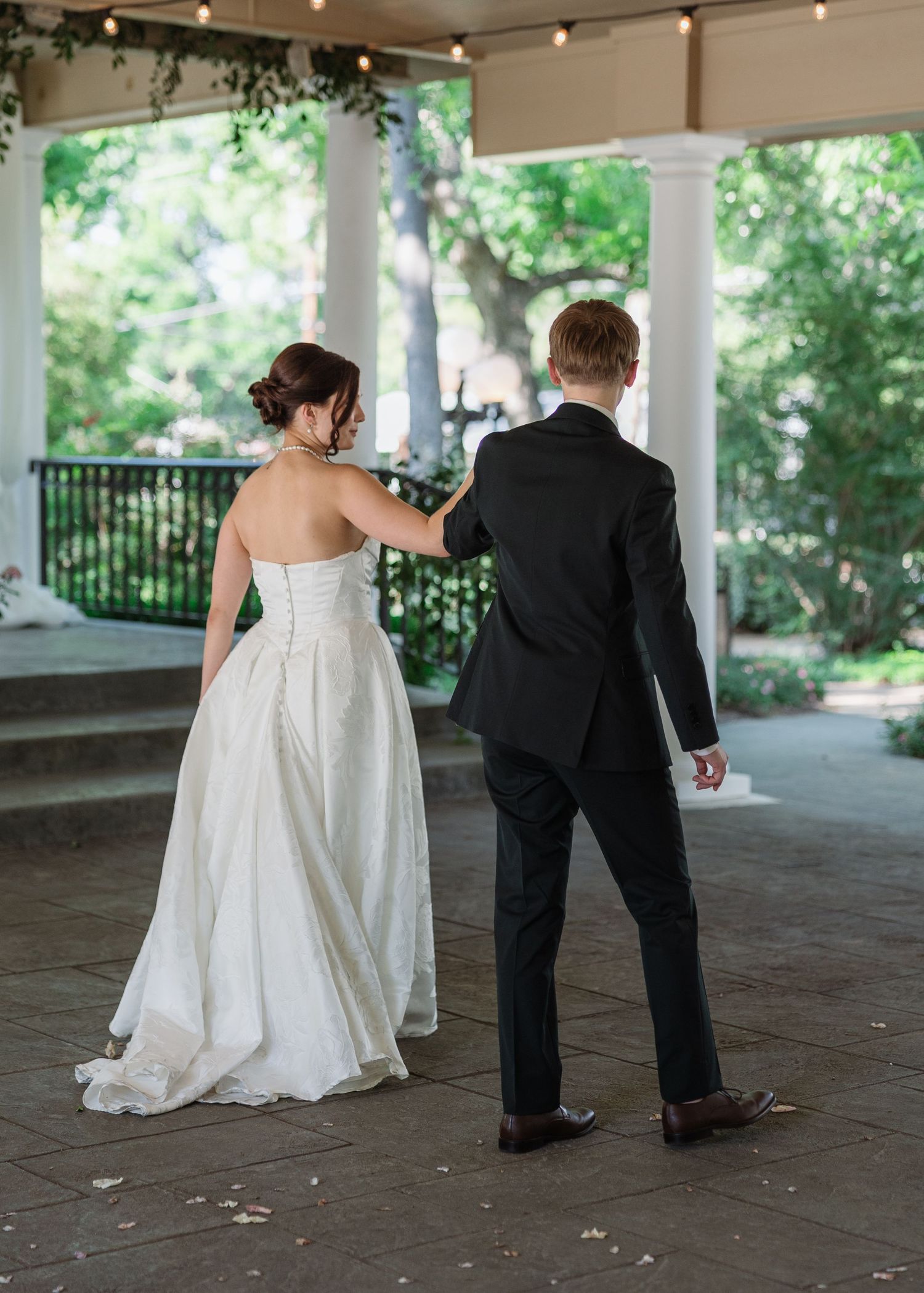 Bride in white strapless gown and groom in black suit walk together under elegant outdoor venue with string lights.