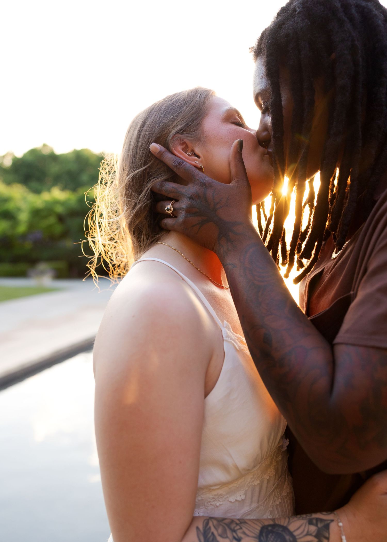 Interracial couple sharing a romantic kiss at golden hour outdoors, woman wearing white dress with engagement ring visible.
