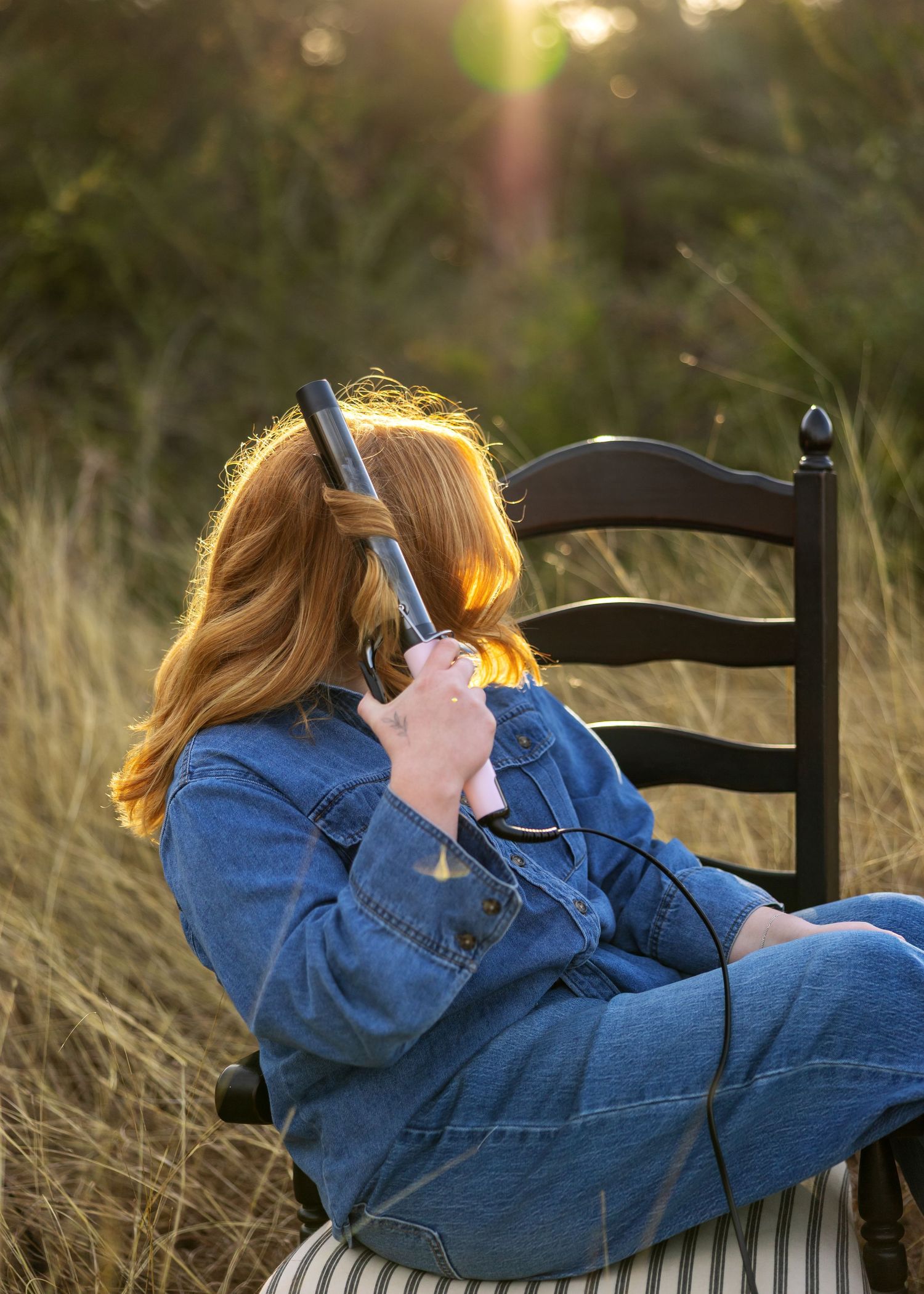 Person in denim jacket lying on black chair outdoors with camera equipment at golden hour.