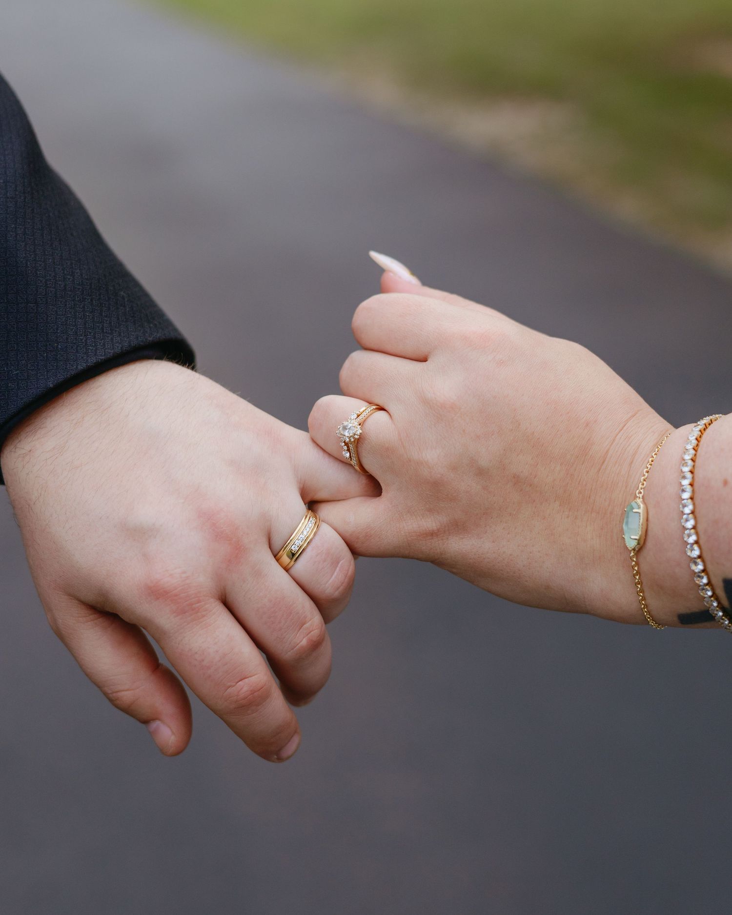 Close-up of couple holding pinkies wearing matching gold wedding bands and elegant bracelets outdoors.