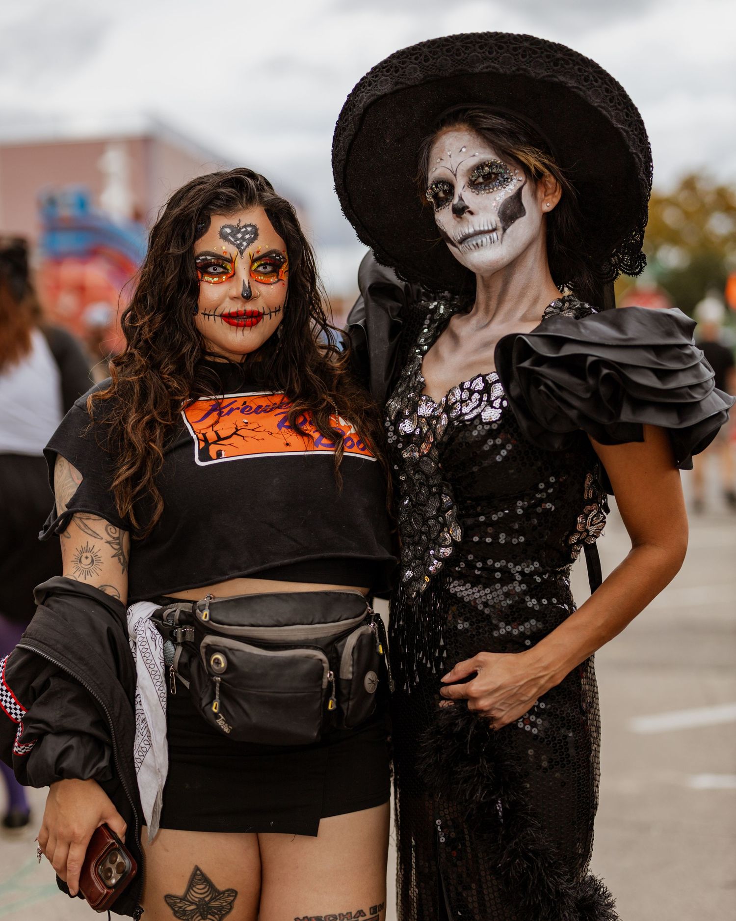Two people in Halloween costumes with Day of the Dead style makeup and black outfits pose together at an outdoor event.