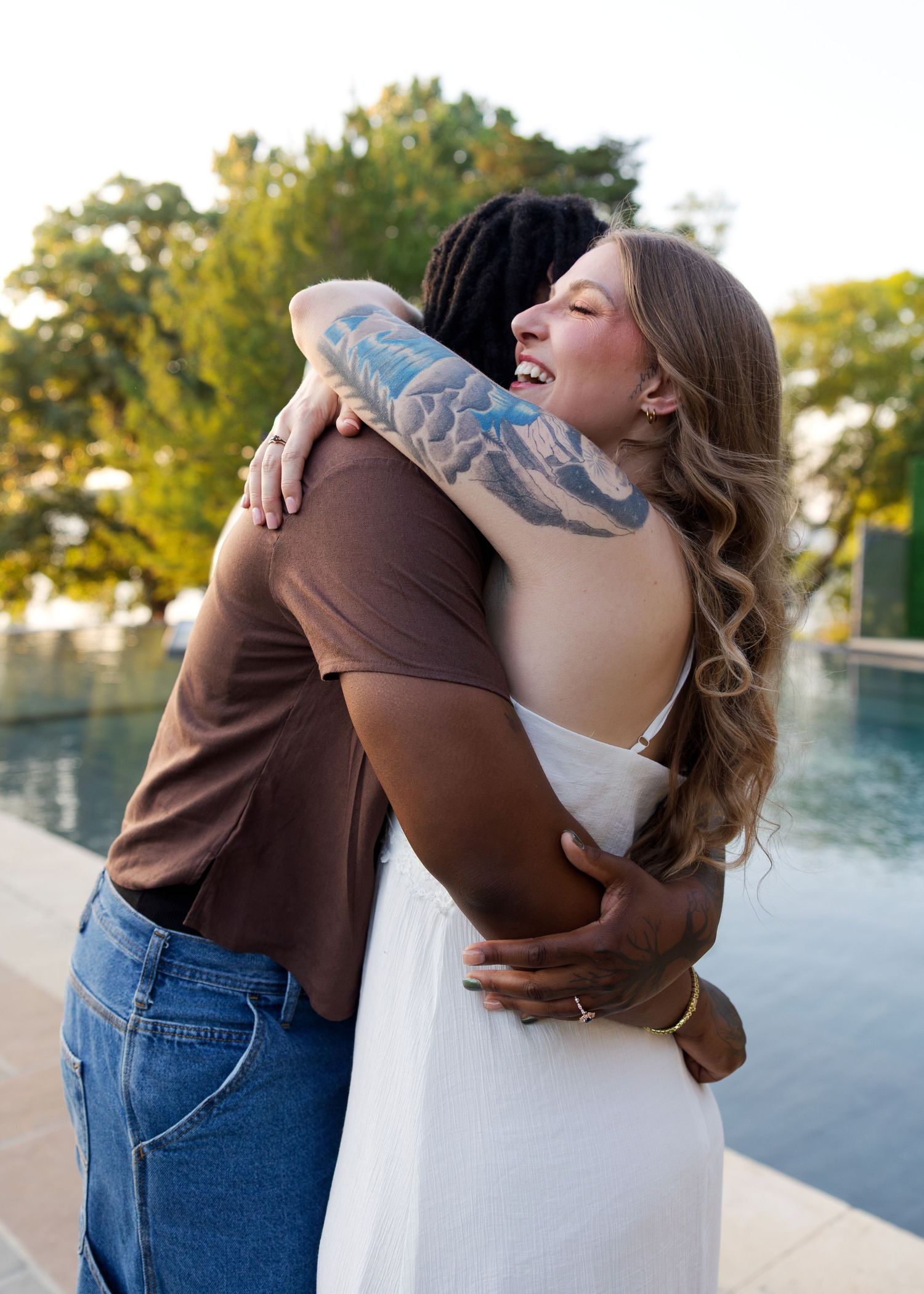 A joyful couple hugging outdoors near a pool, woman in white dress with a tattoo sleeve smiling.