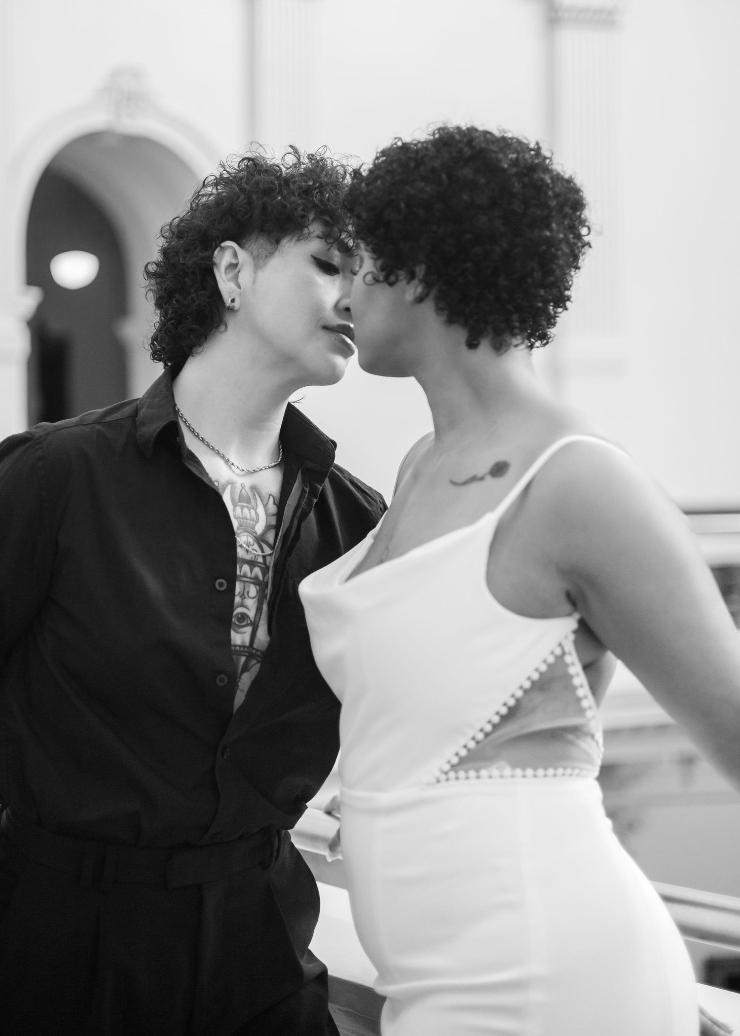 Two people with curly hair share a romantic moment, one in black outfit, other in white dress, black and white photo.
