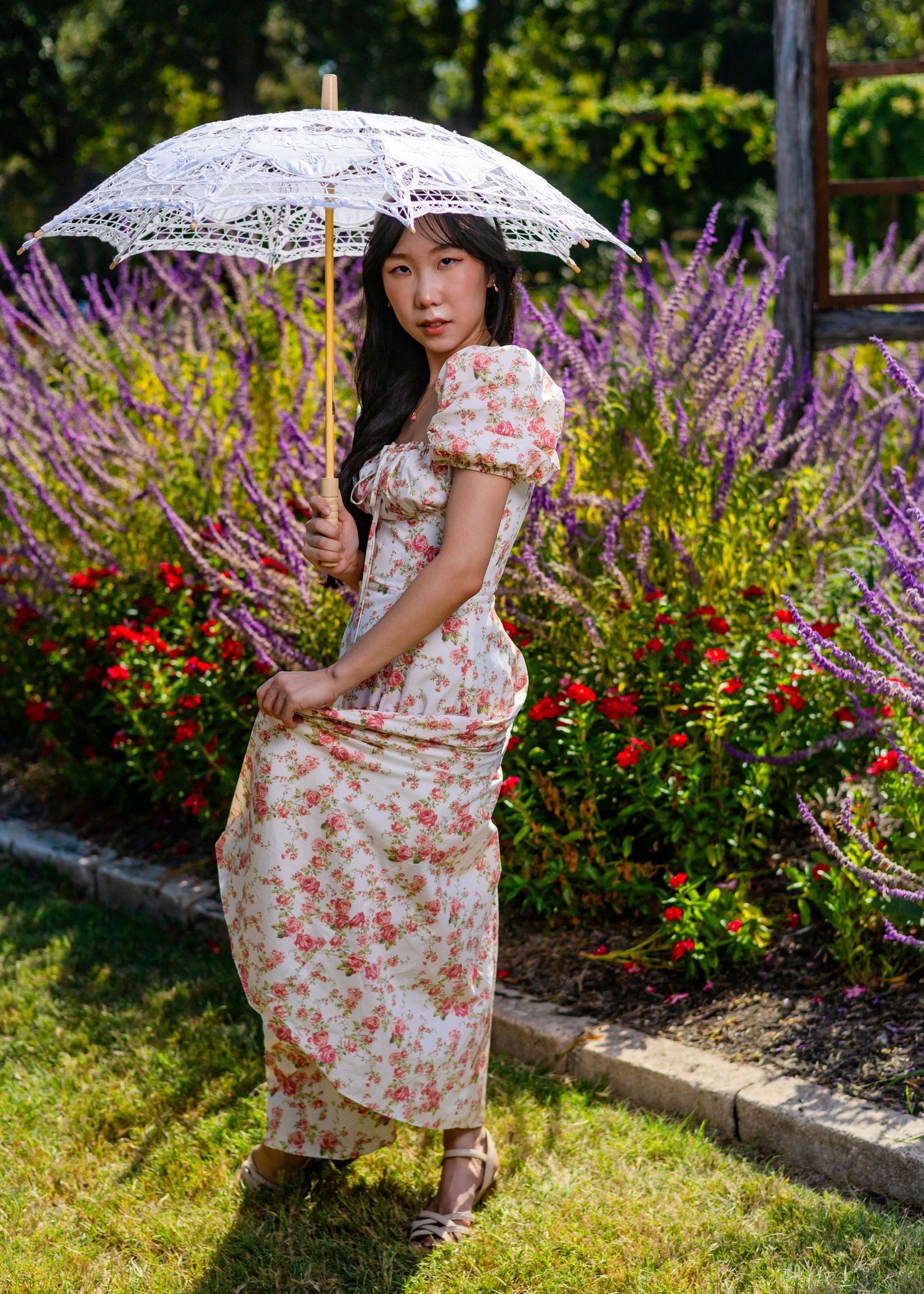 Garden photoshoot featuring white lace parasol and floral dress amid blooming flowers.