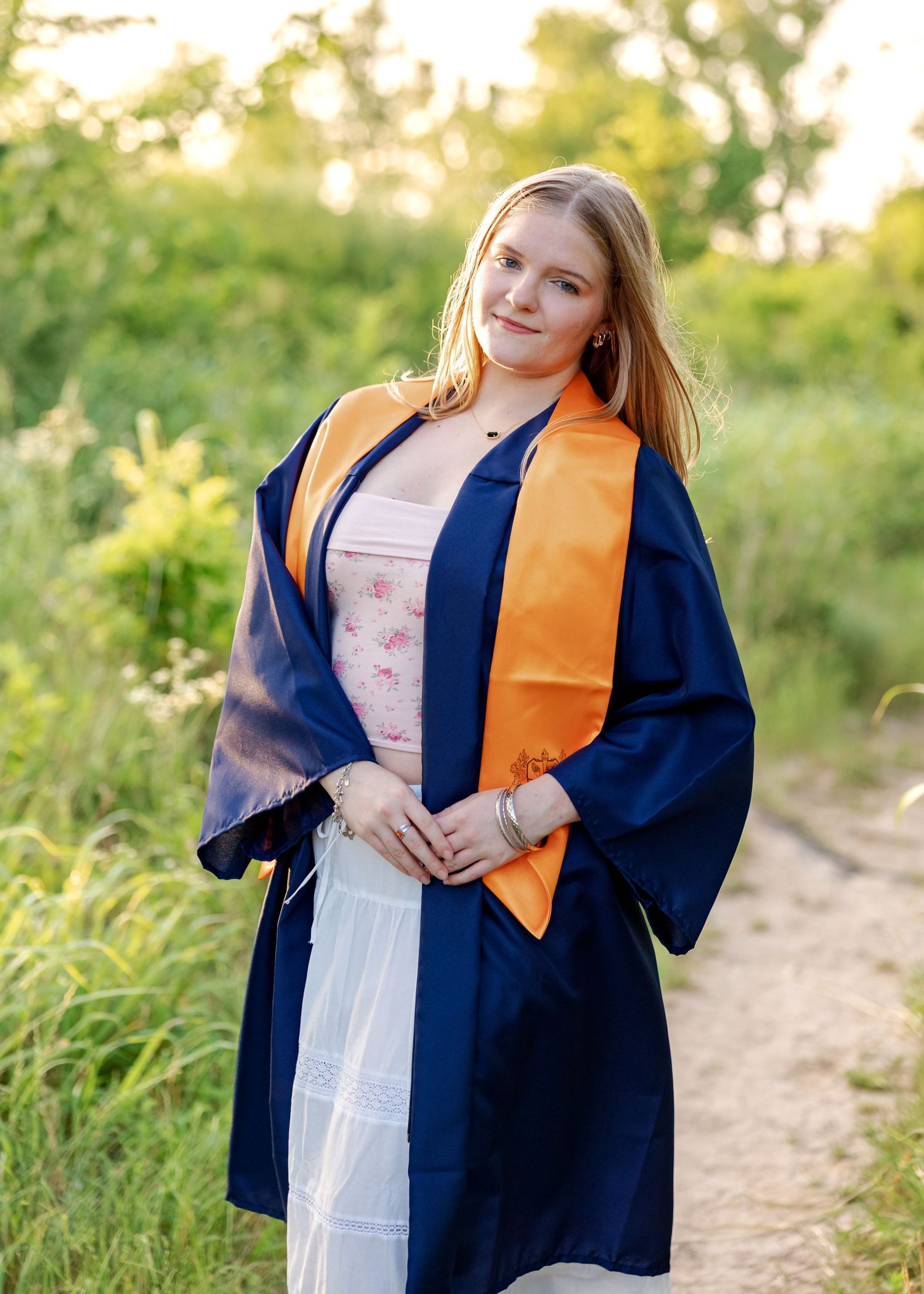 Young woman in navy blue graduation gown and orange stole poses outdoors on a sunny day.