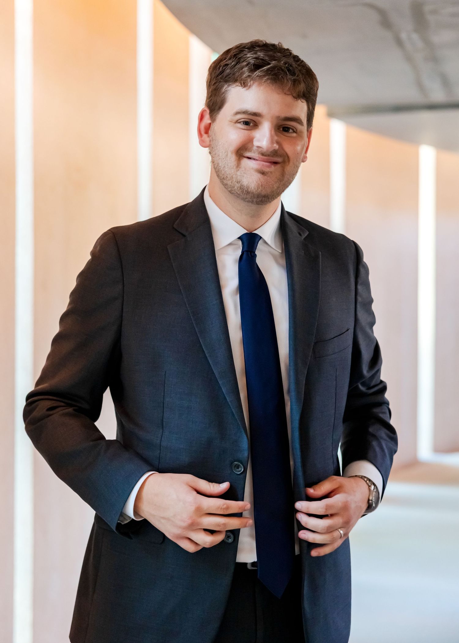 Young professional man in dark suit and navy tie smiling confidently in modern corridor.