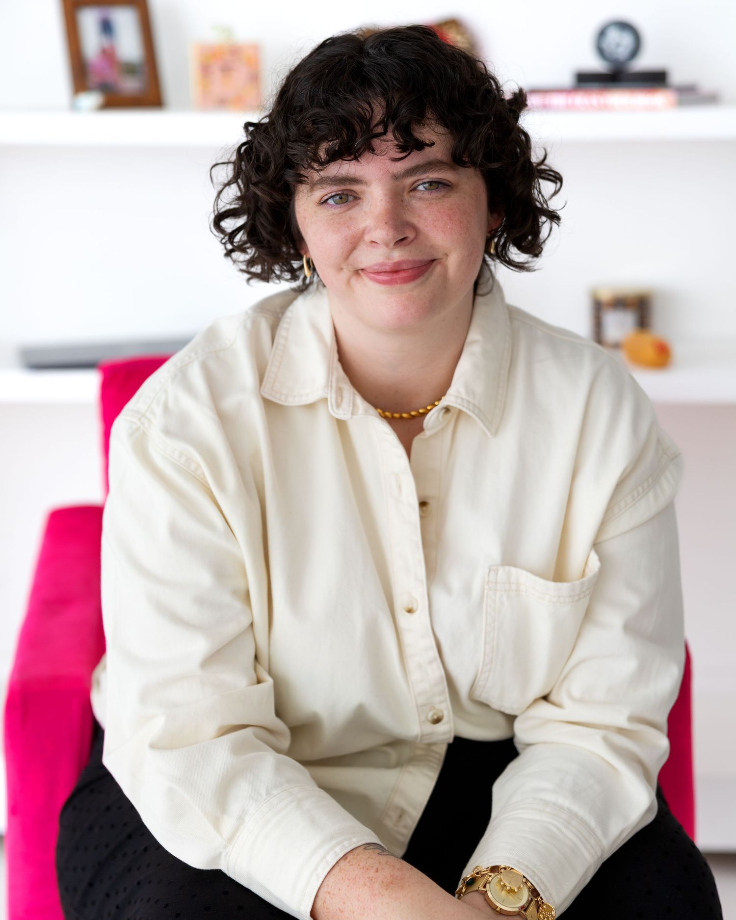 A person wearing a cream colored button-down shirt sits casually against a white background with pink accents.