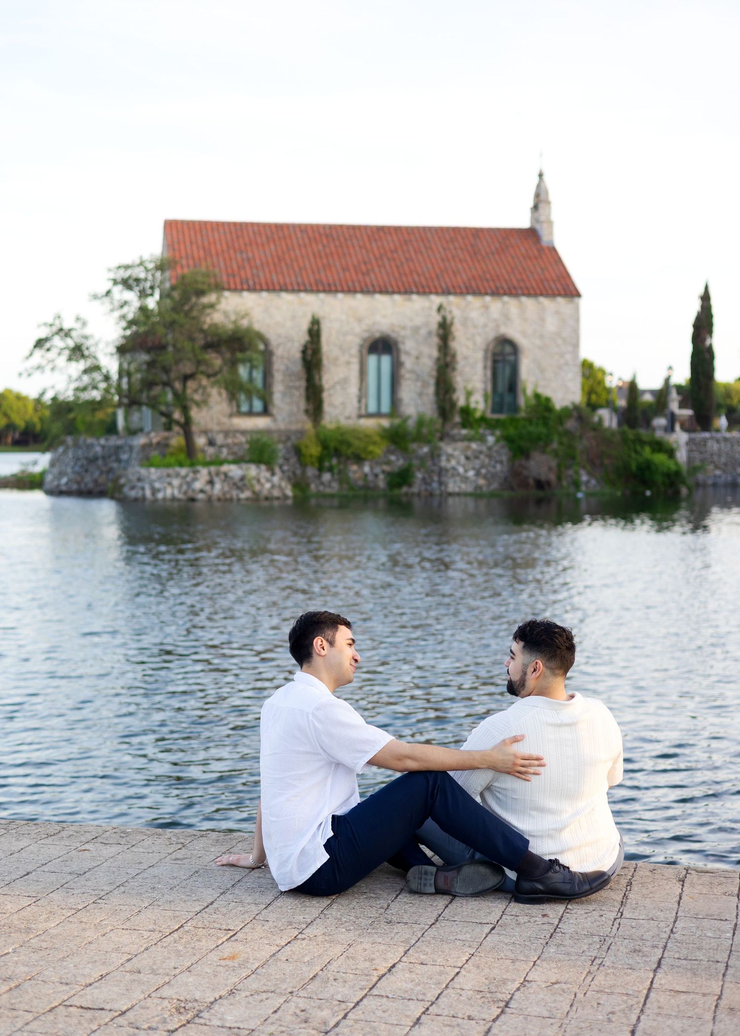 Two men sitting together by a lakeside, facing each other with a historic stone chapel on an island in the background.