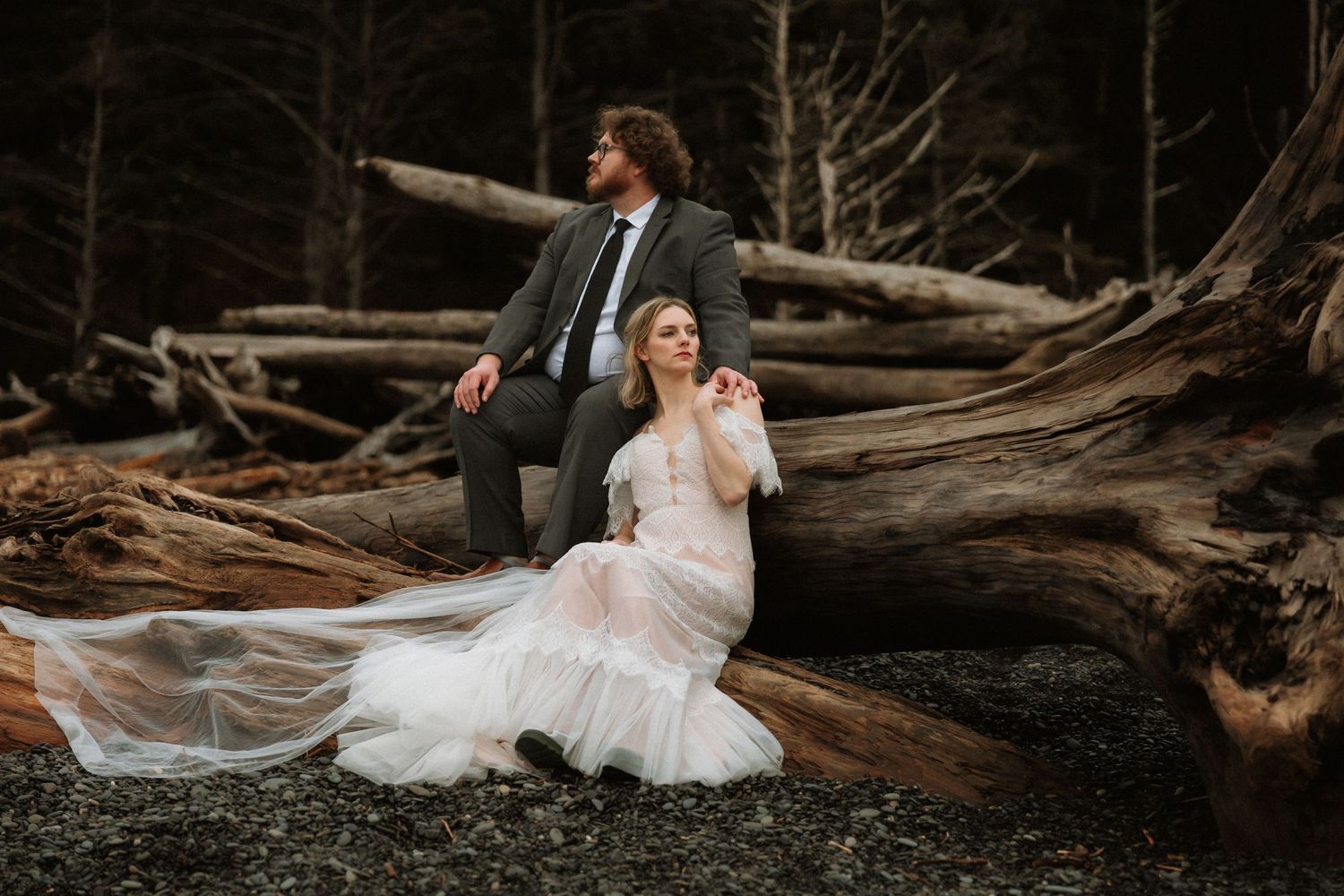 Bride in flowing white gown sits on driftwood with groom in gray suit, surrounded by dark forest trees.