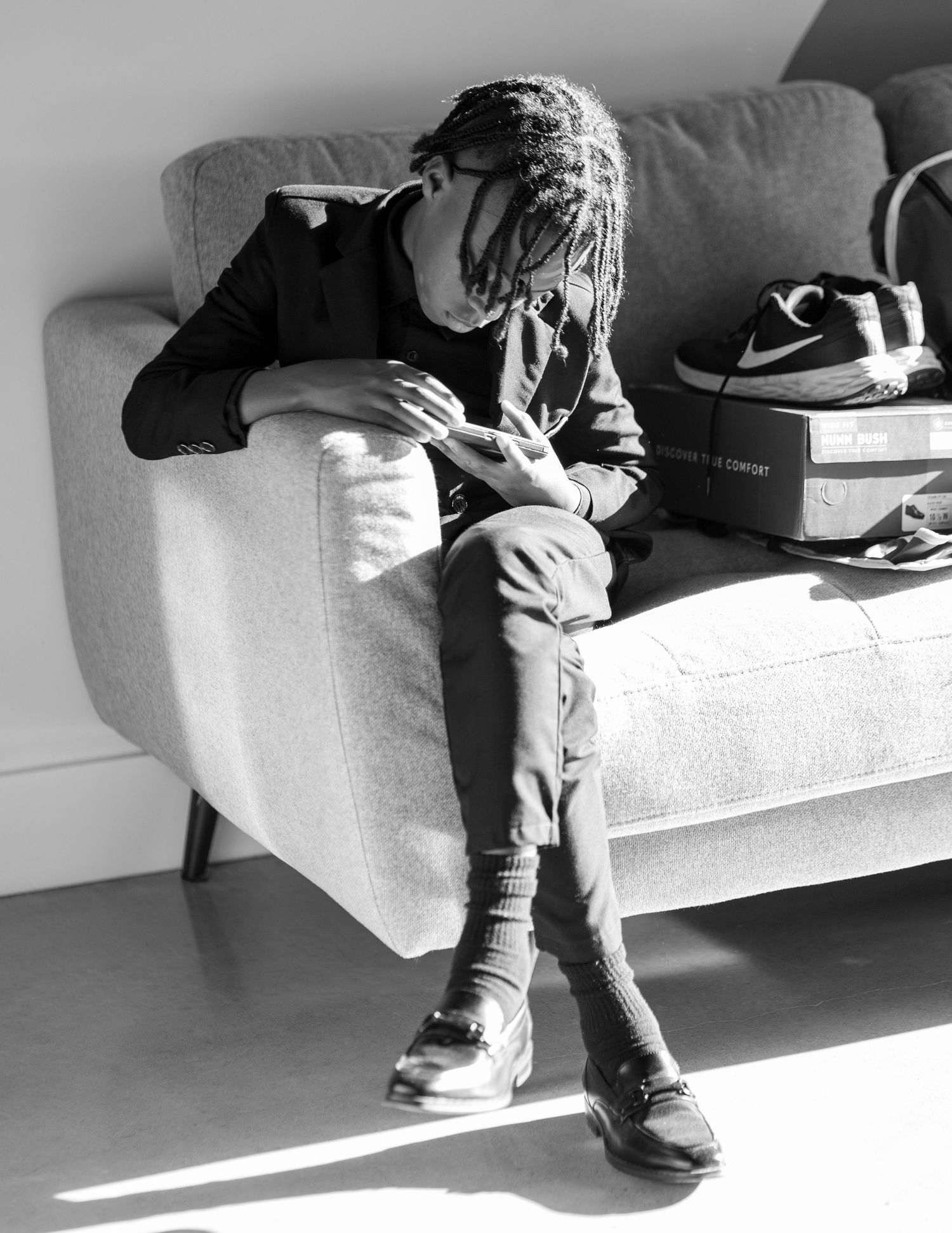 Black and white photo of a person with dreadlocks sitting on a gray sofa, looking at their phone.