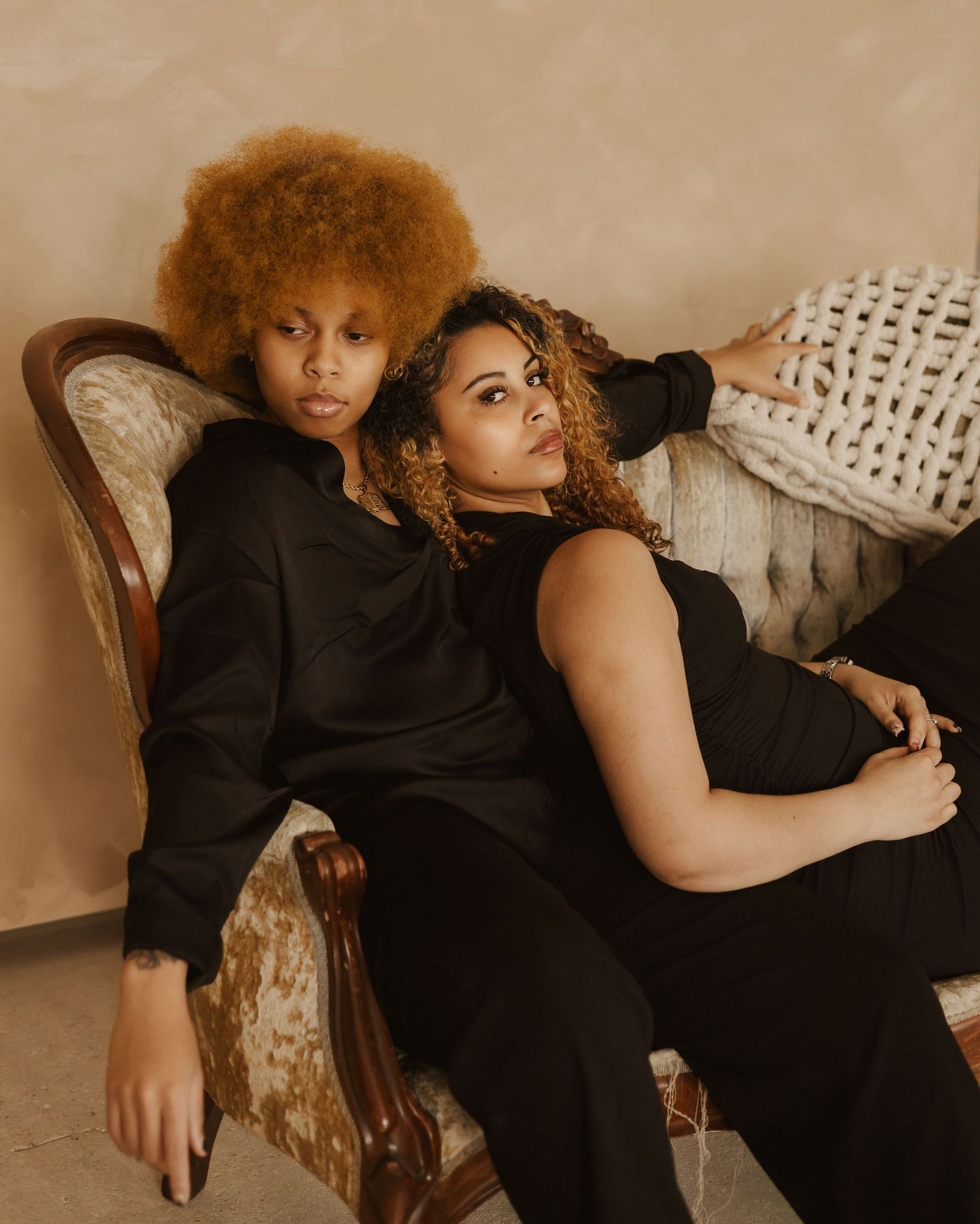 Two people in dark clothing pose together on a beige couch in a warm-toned studio setting.
