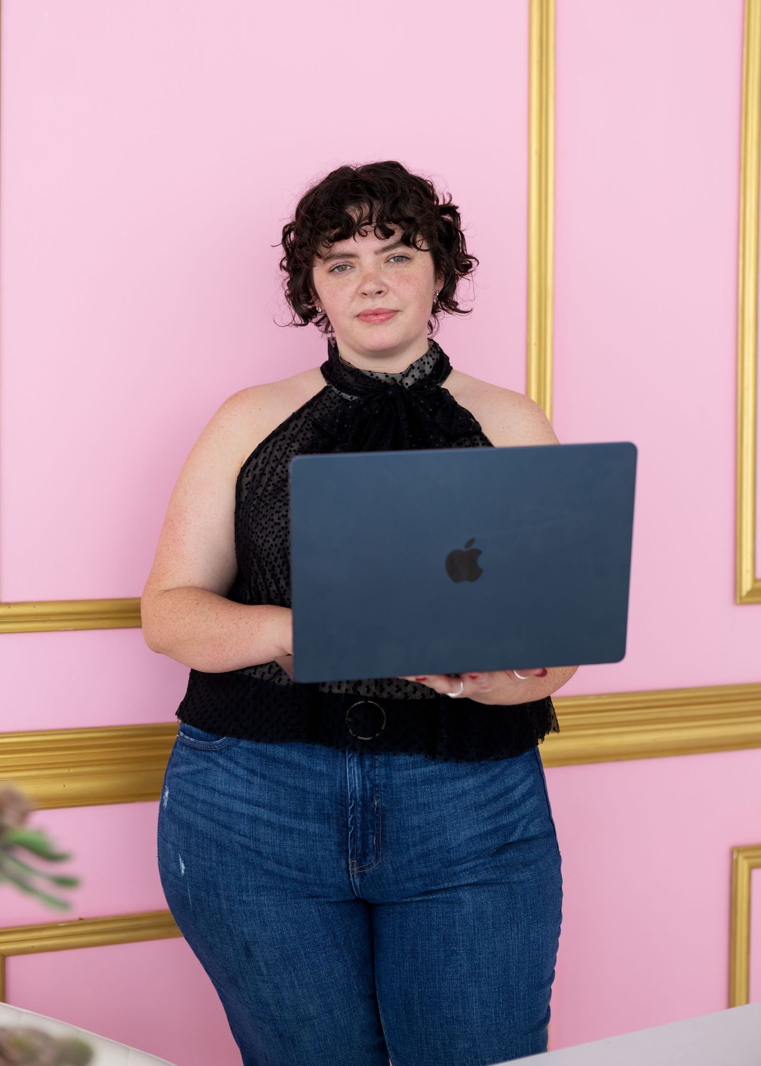 Person in black top using laptop while seated against pink wall with gold trim molding.