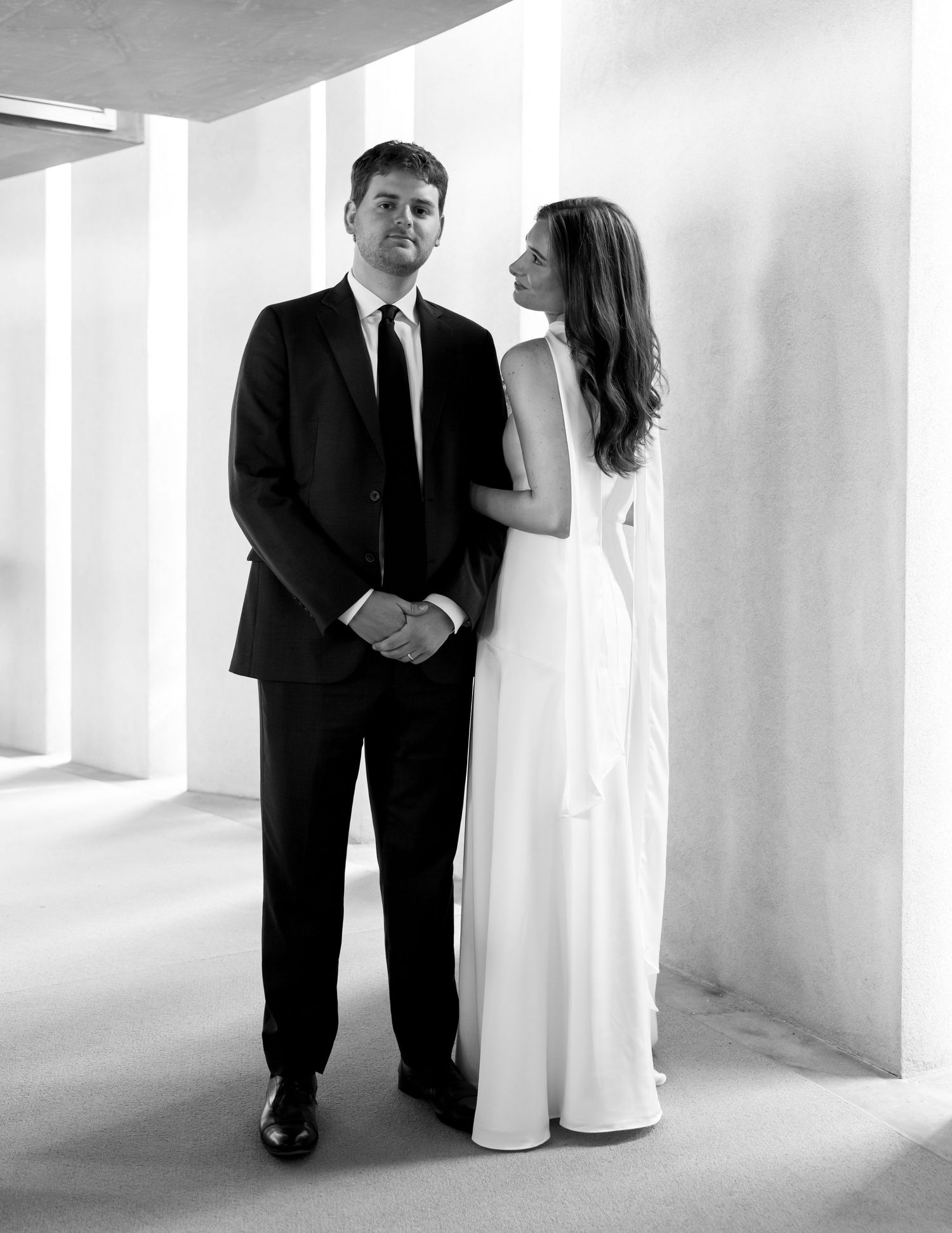 Bride in white gown and groom in black suit pose together in a modern architectural setting.