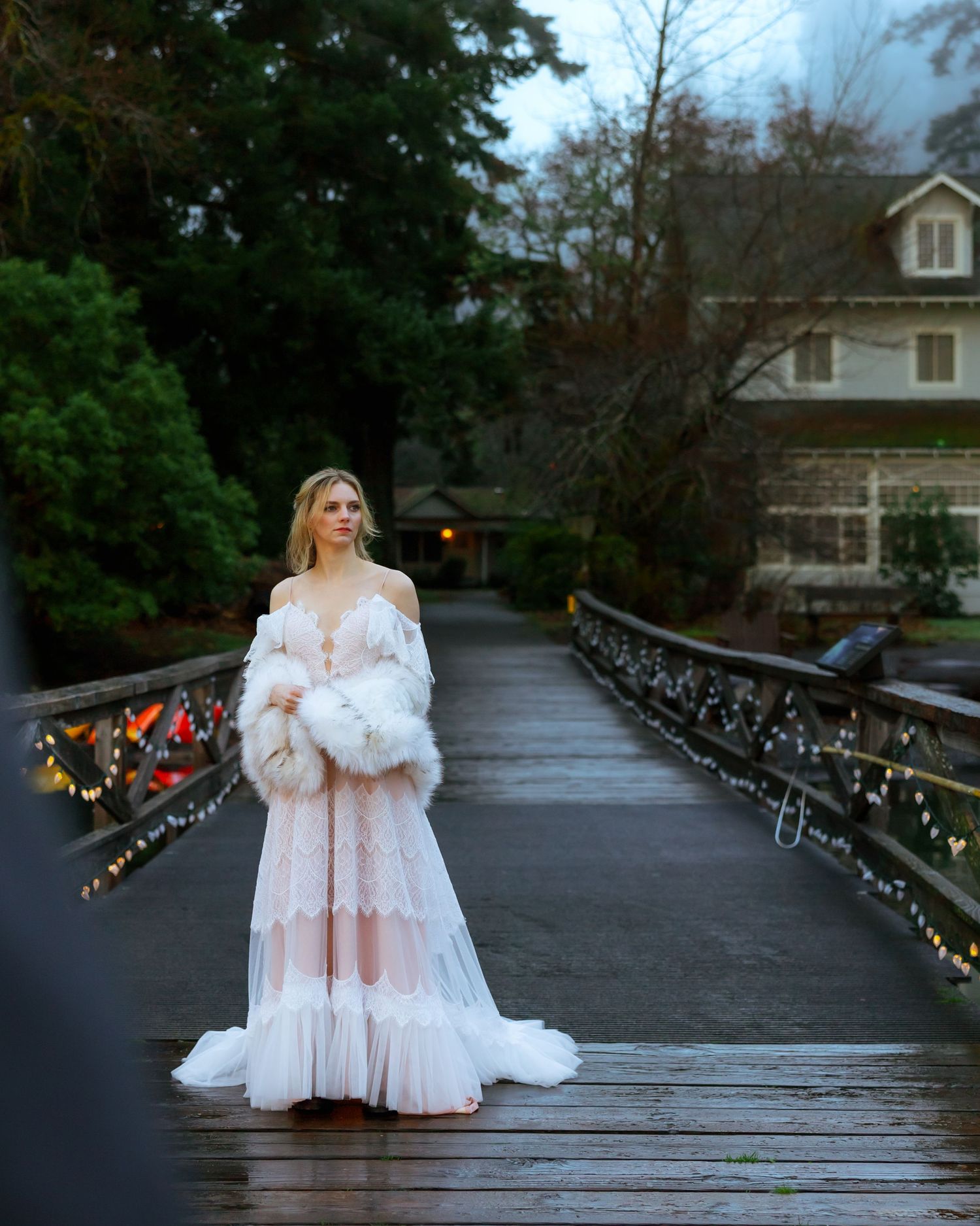 Bride in flowing lace gown and fur stole stands on wooden bridge surrounded by lush greenery.