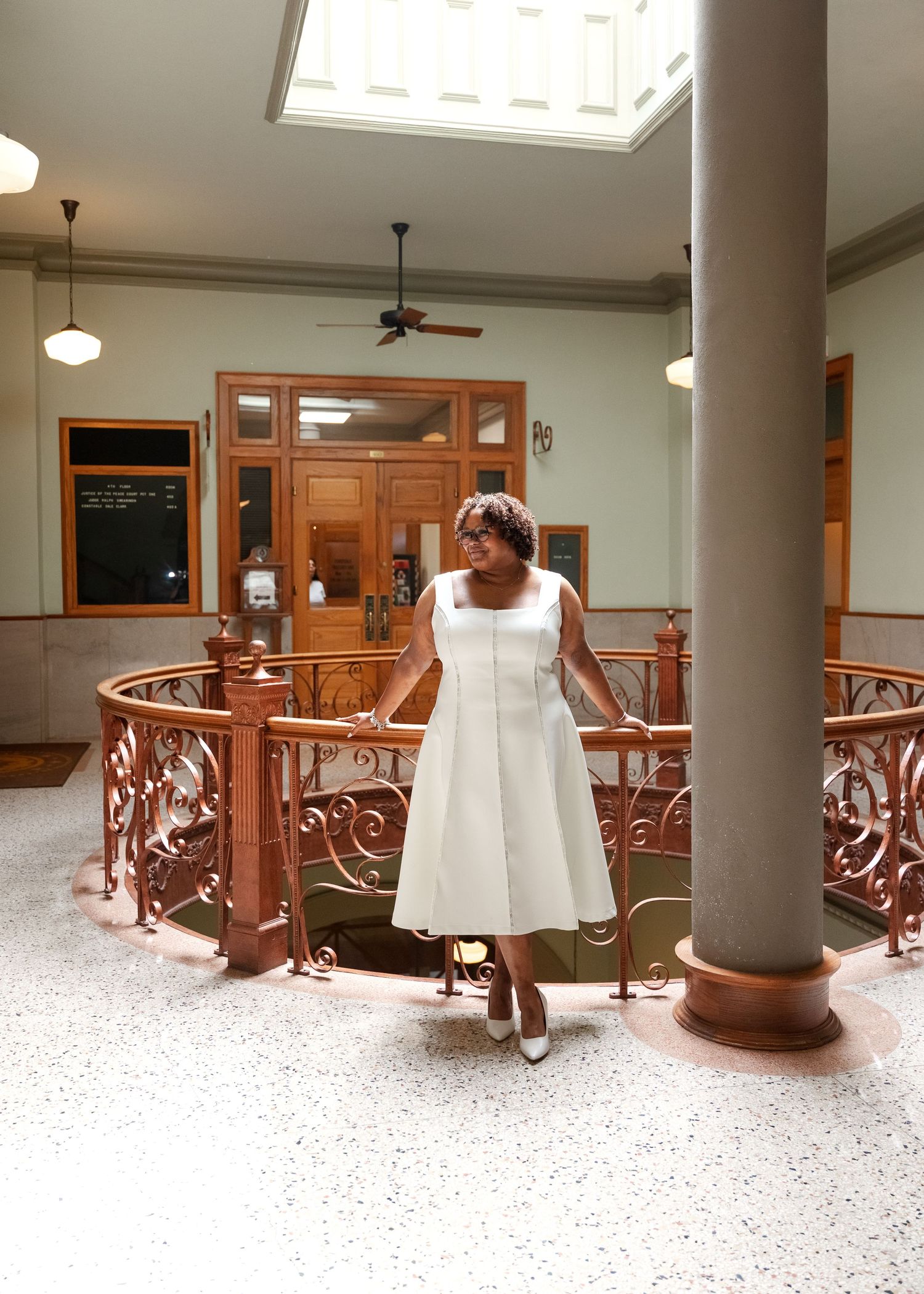 Woman in white midi dress stands on ornate railing balcony inside elegant historic building with marble floors.