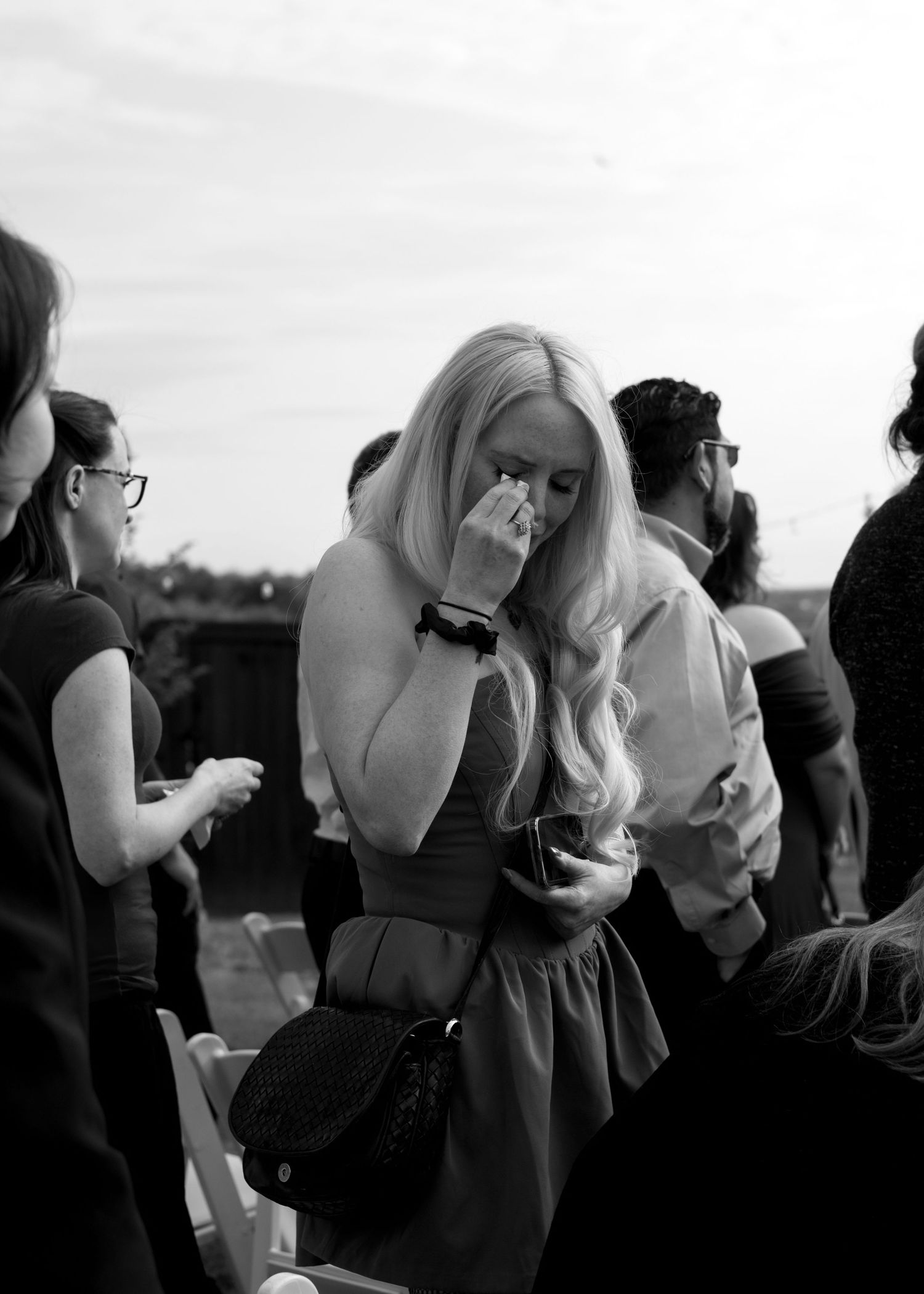 Black and white photo of emotional blonde woman wiping tears at an outdoor wedding ceremony.