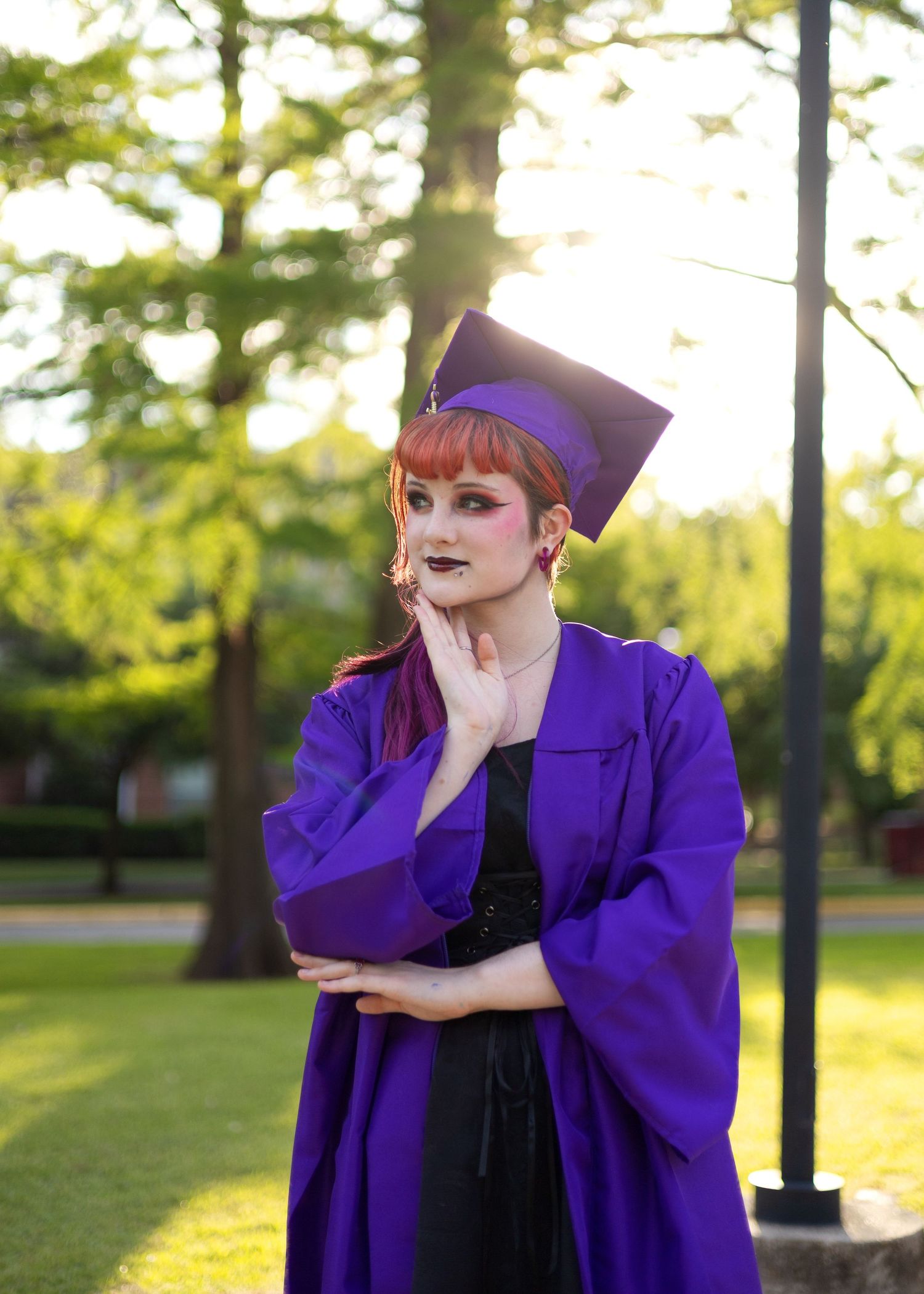A young woman in a purple graduation cap and gown poses outdoors among green trees in a park setting.