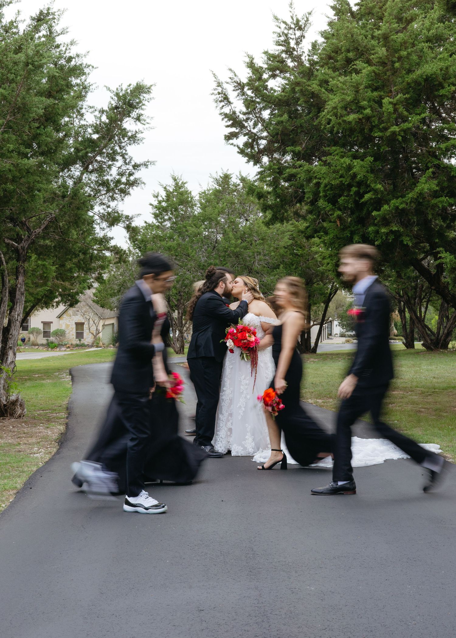 Bride and groom share a kiss on a tree-lined path while blurred wedding party members walk past in black attire.