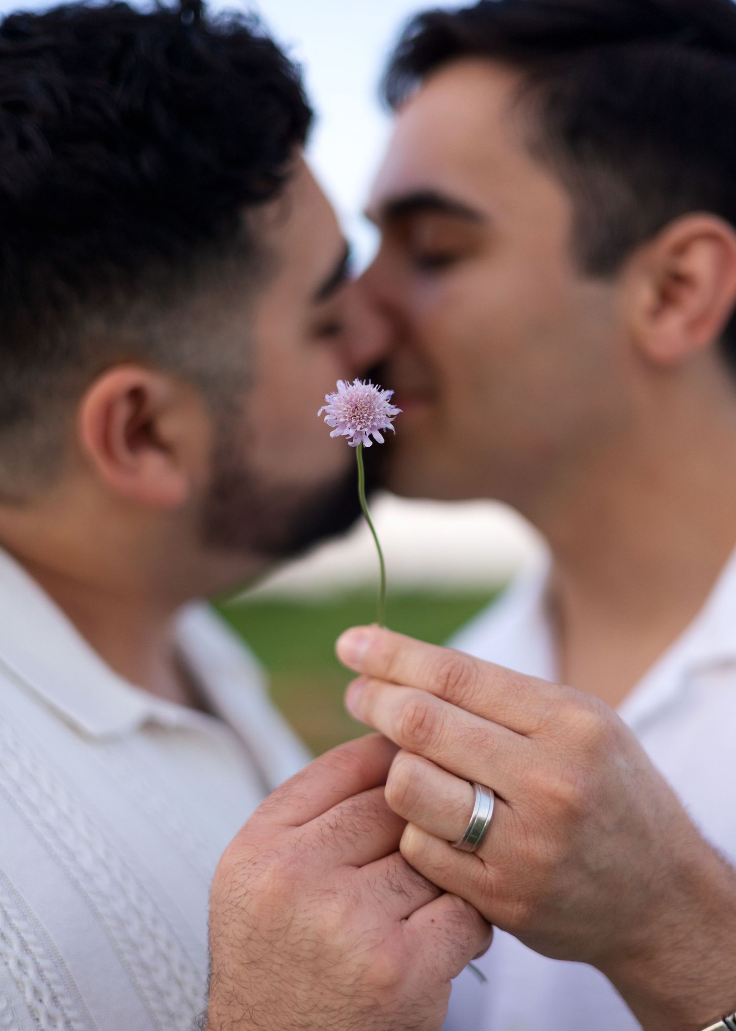 Two men sharing a romantic kiss while holding a delicate purple flower, one wearing a silver wedding band.