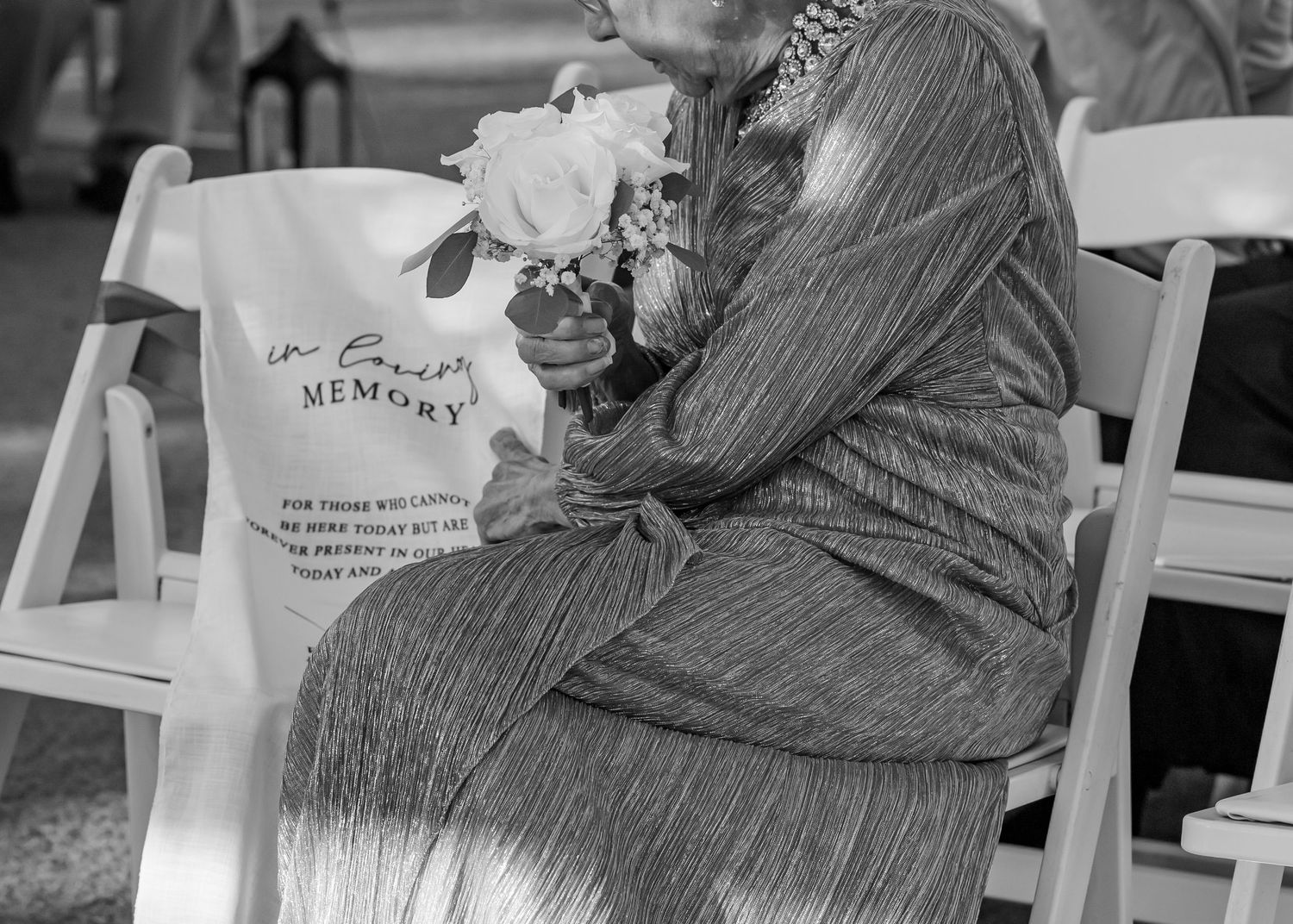 Elderly woman seated at memorial service holding white flowers beside an 'In Loving Memory' bag.