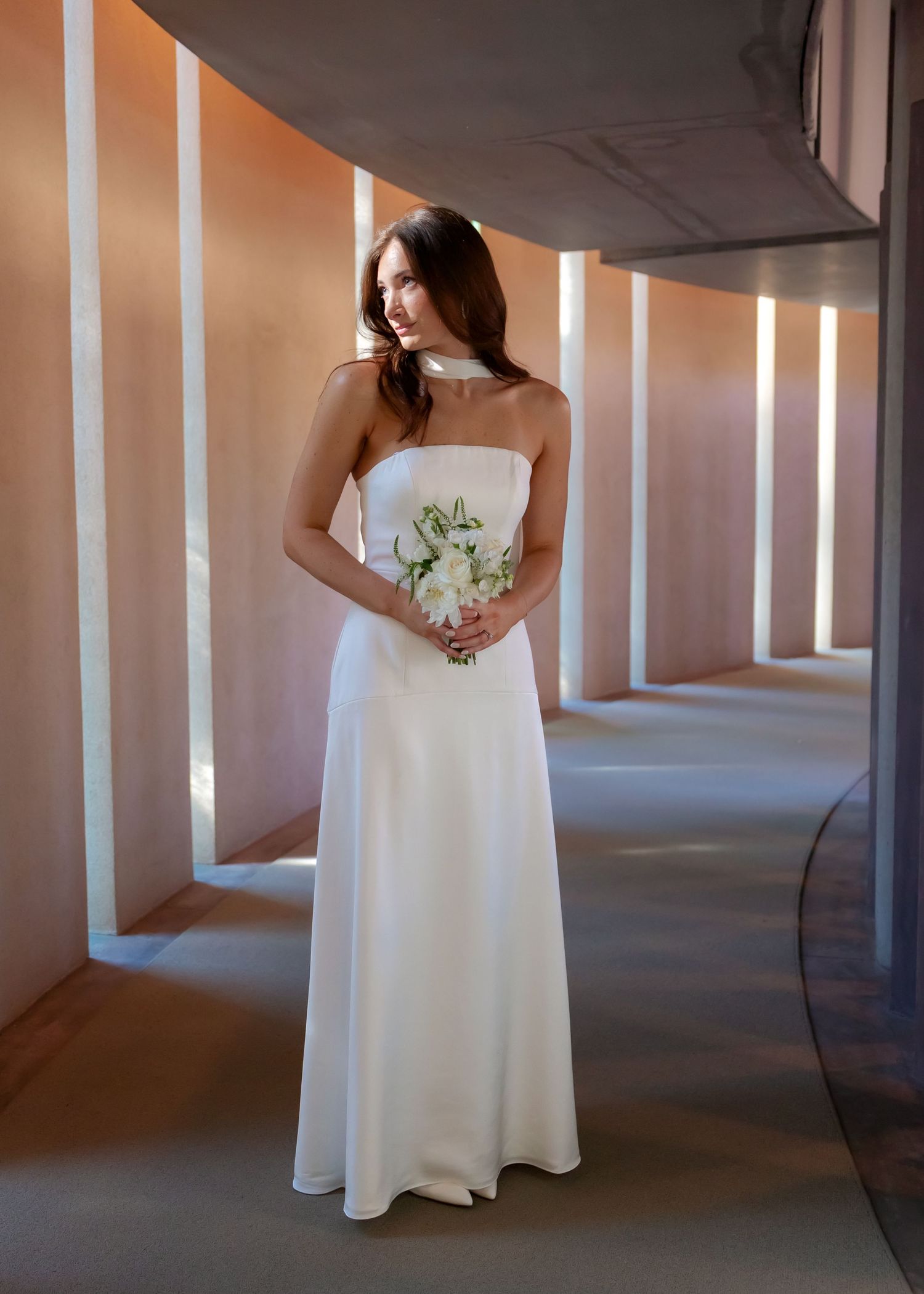 Elegant bride in strapless white wedding dress holding bouquet in warmly lit curved corridor.