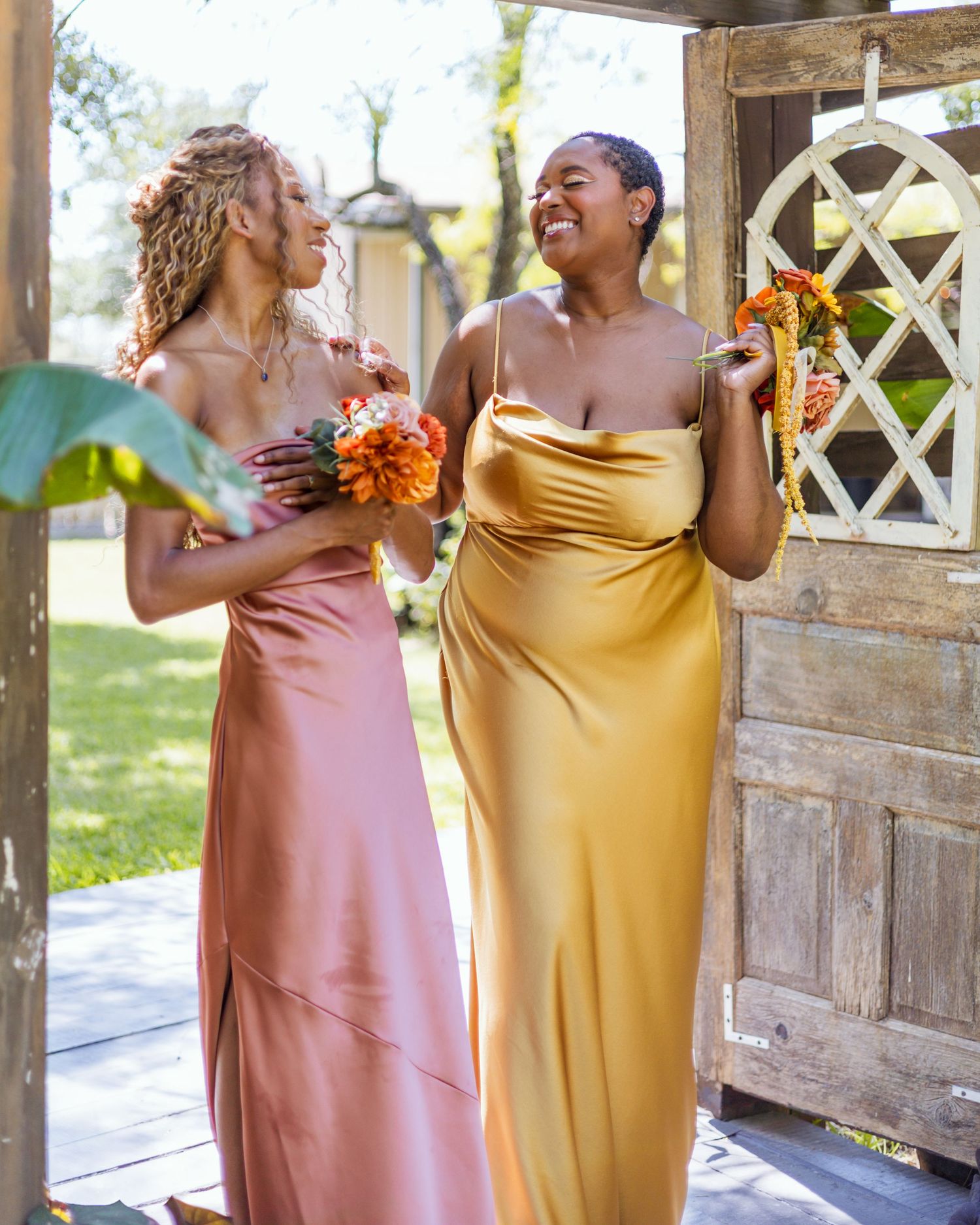 Two bridesmaids in satin gowns holding orange bouquets laugh joyfully at a rustic wooden doorway outdoors.