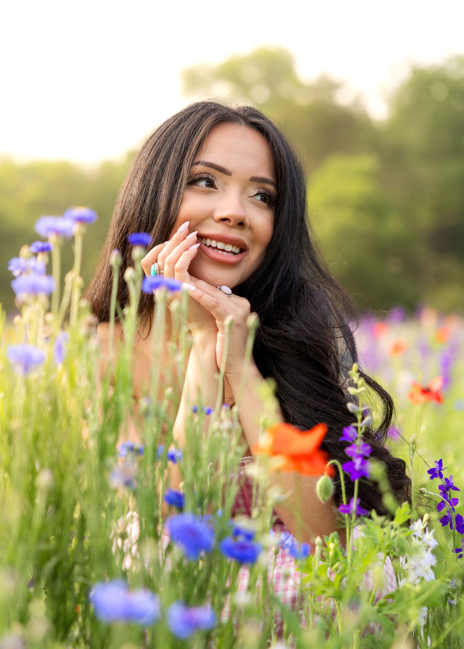 Smiling young woman with long dark hair lying in a colorful wildflower field with blue and red blooms.