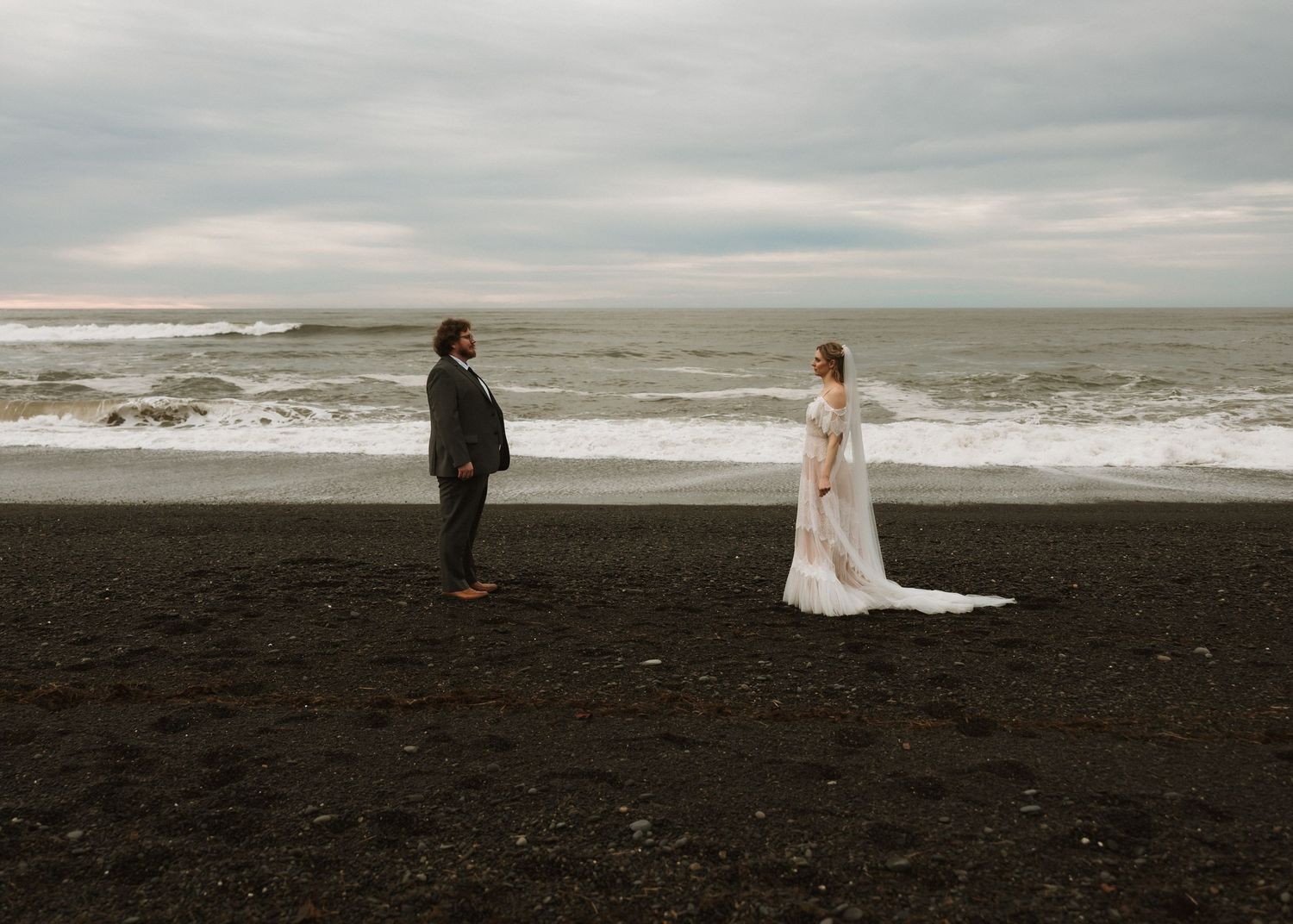 Bride and groom stand apart on a black sand beach with dramatic ocean waves under a cloudy sky.
