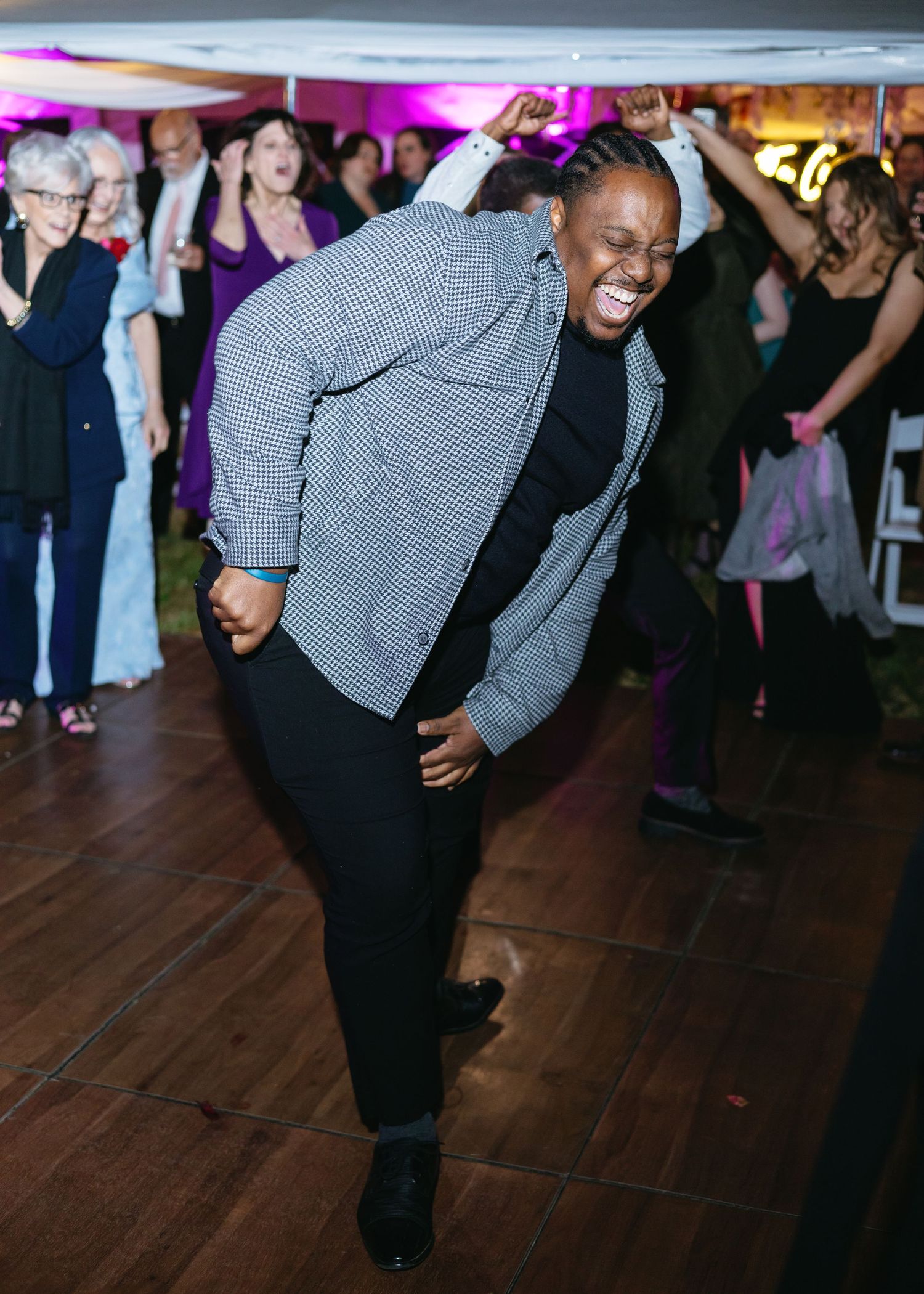 Joyful wedding guest in checkered jacket dances energetically on the reception dance floor with crowd behind.