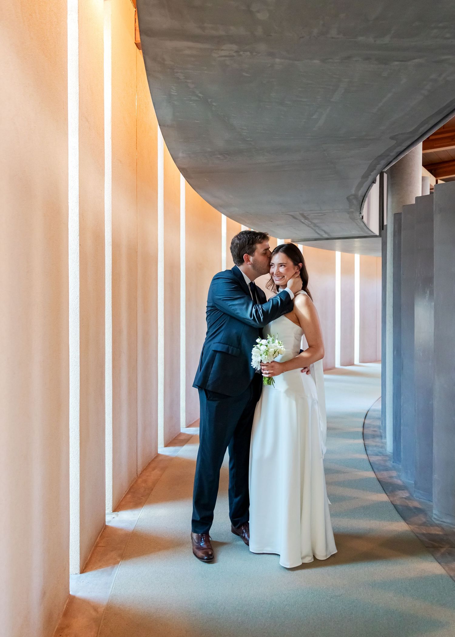 Bride and groom share a romantic kiss in a modern curved hallway with elegant lighting on their wedding day.