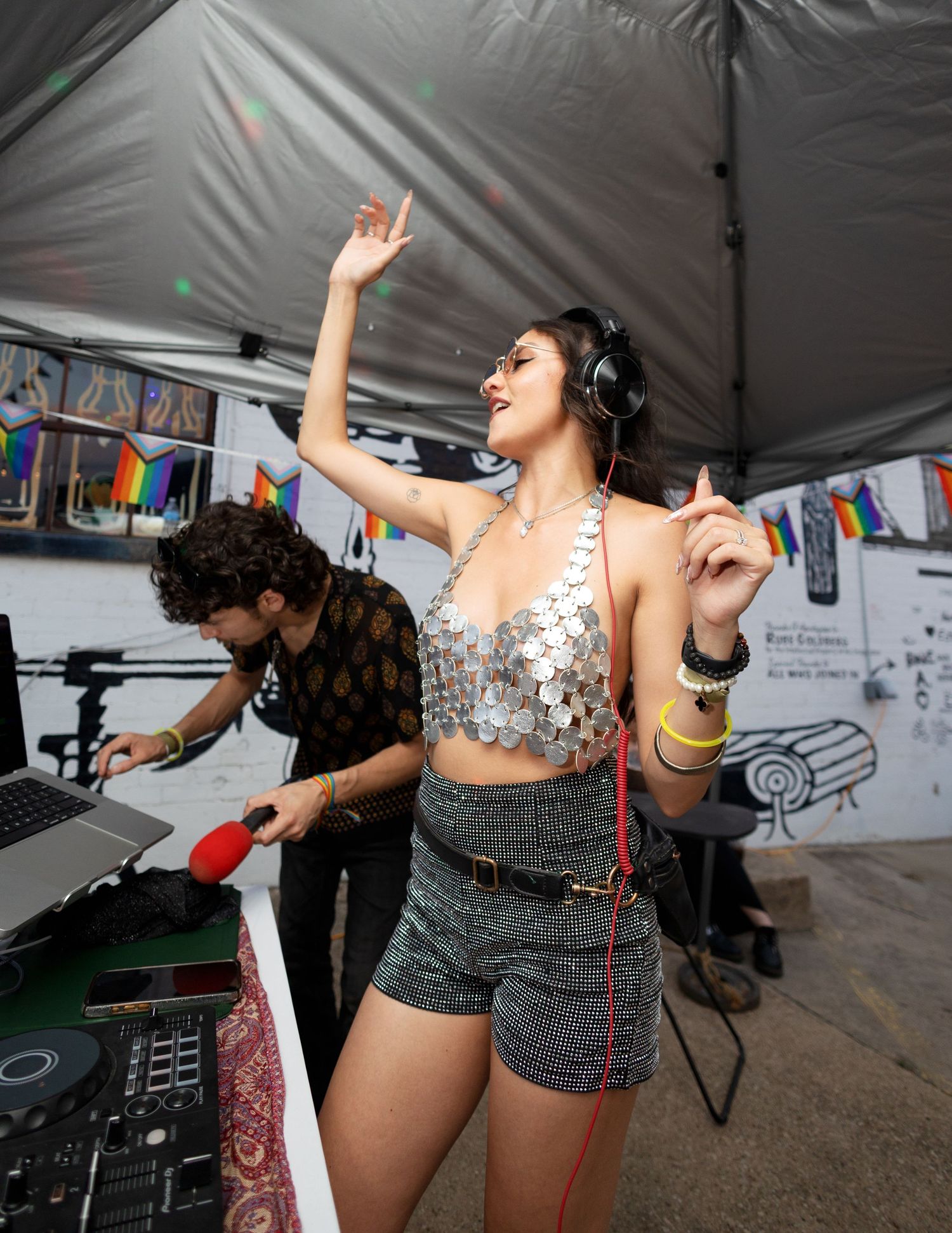 Woman in silver sequin top dancing at outdoor DJ event with pride flags and DJ mixing in background.