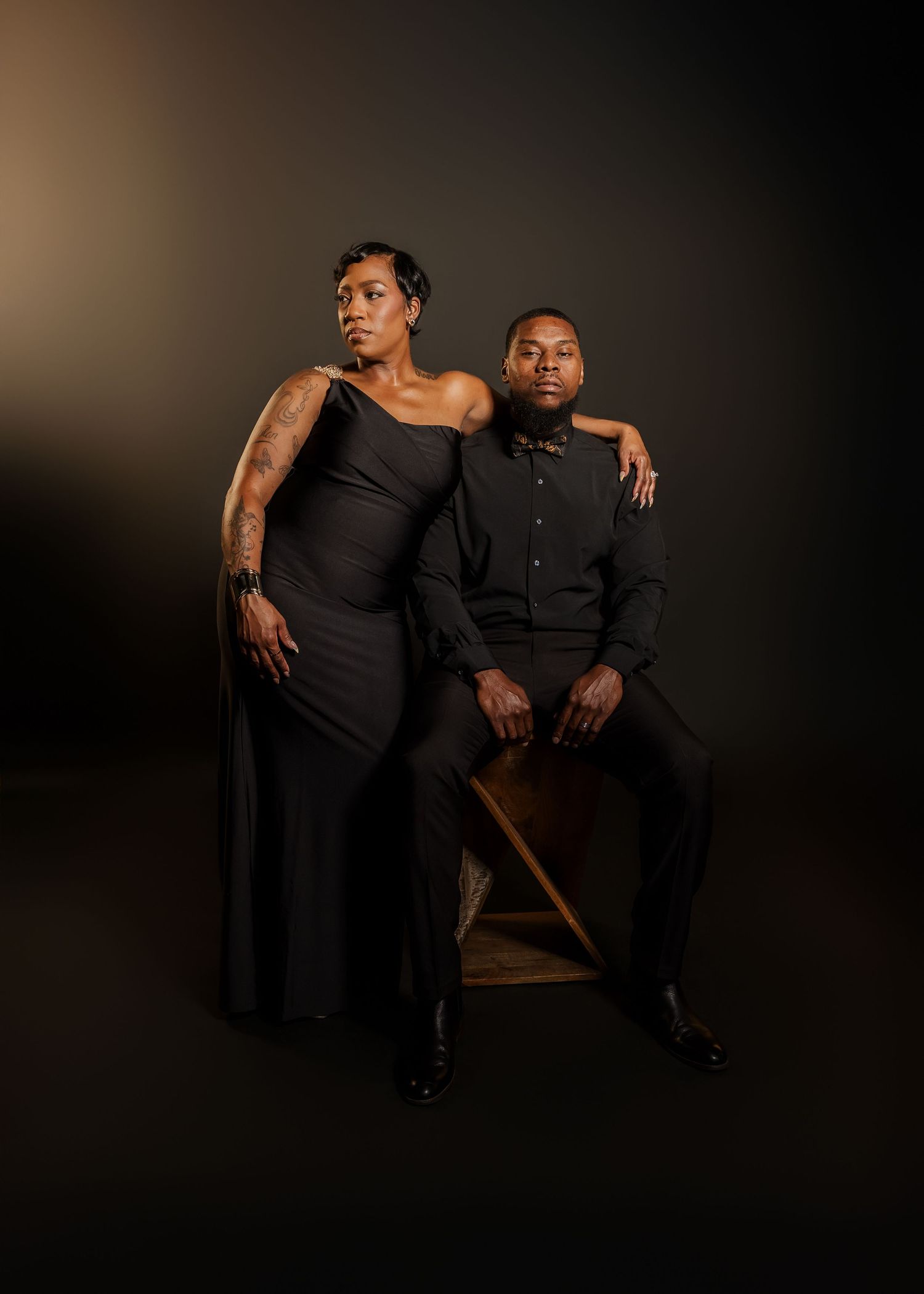Two people in elegant black outfits pose dramatically in a studio with warm lighting.
