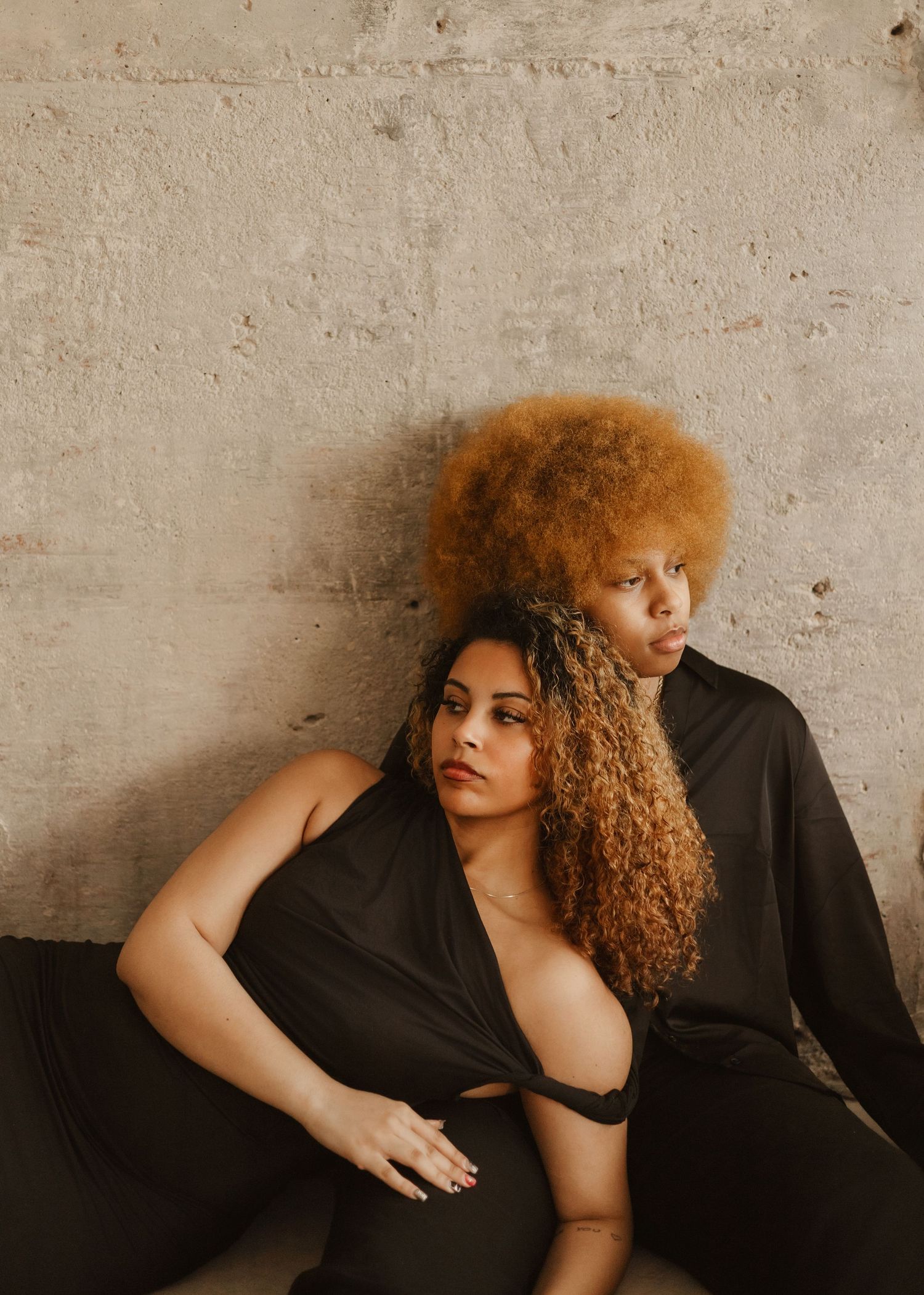 Two models with natural curly hair pose together against a concrete wall wearing black outfits in moody lighting.