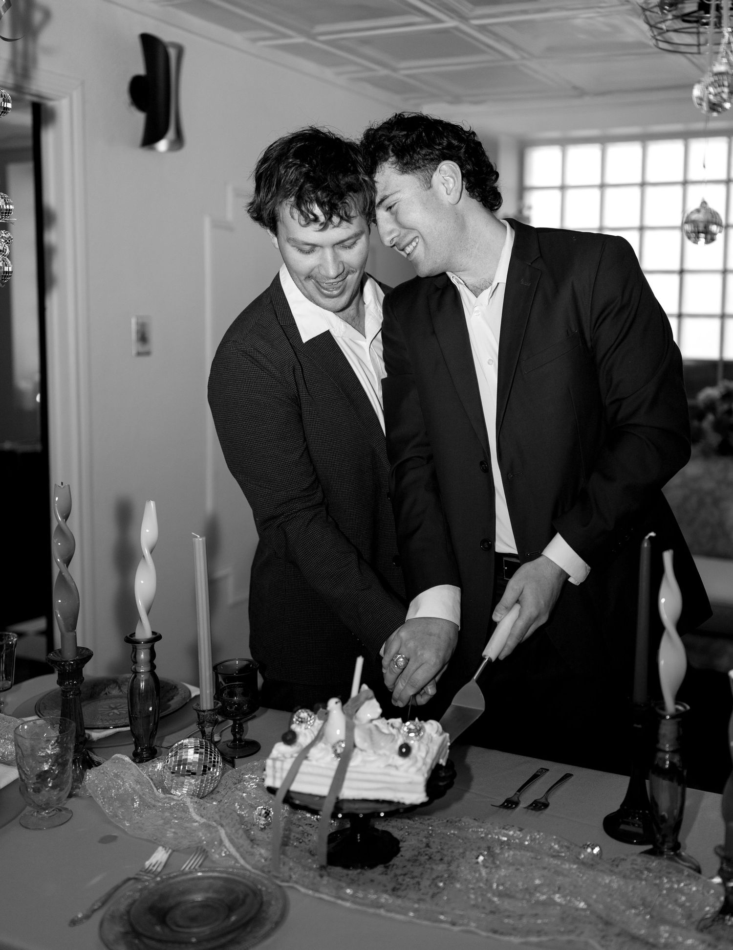 Two men in dark suits cutting a wedding cake together at a reception table in black and white photo.