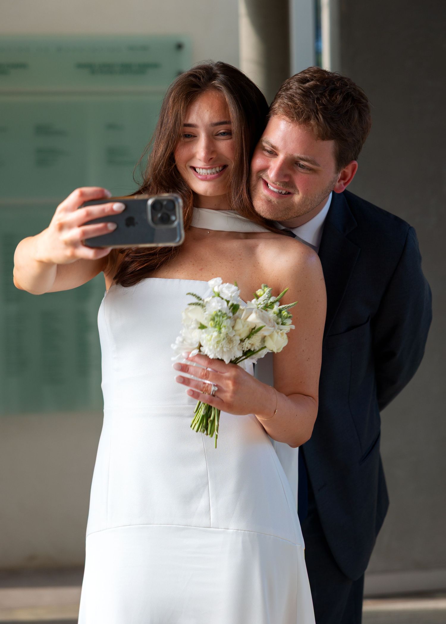 Bride in white dress holds bouquet and takes selfie with groom in navy suit on their wedding day.