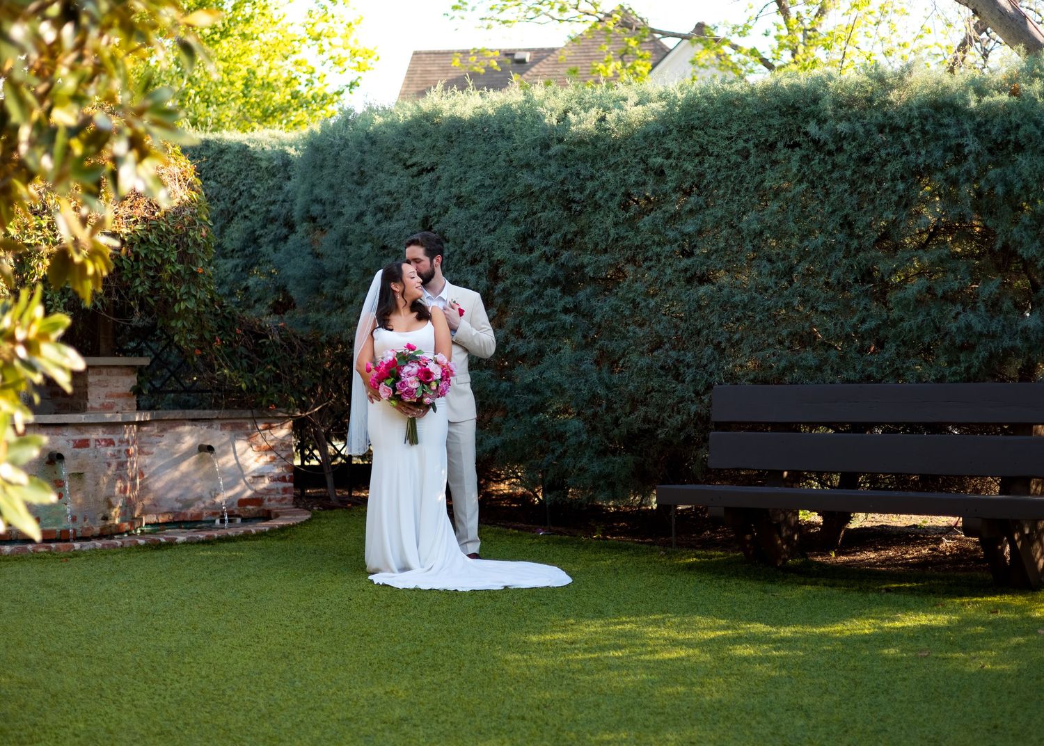 Bride and groom embracing in a lush garden with a white dog, dark hedge backdrop, and wooden bench nearby.