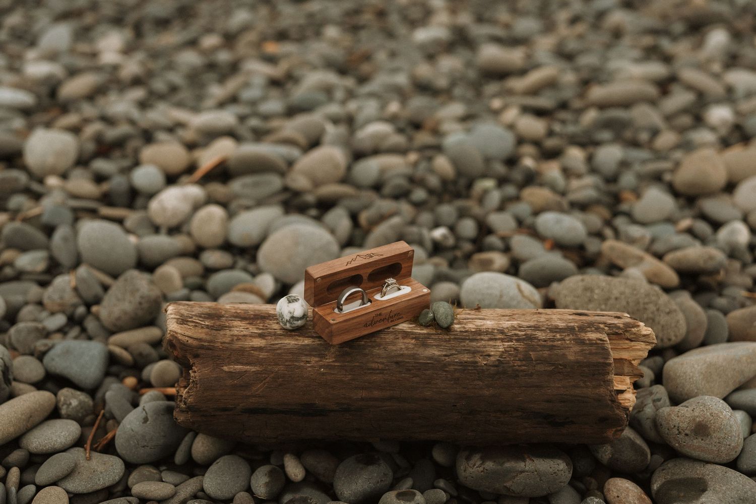 Wooden ring box open on driftwood surrounded by smooth pebbles on a rocky beach, displaying wedding rings.