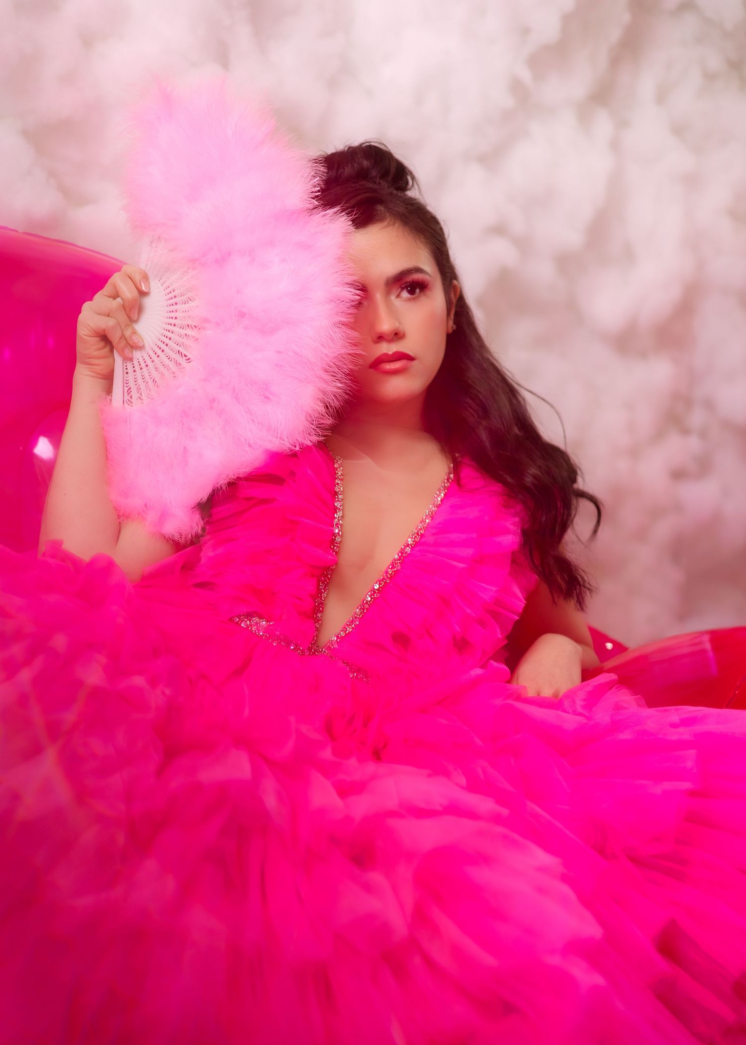 Glamorous portrait in hot pink tulle gown with matching feather fan against cloudy backdrop.