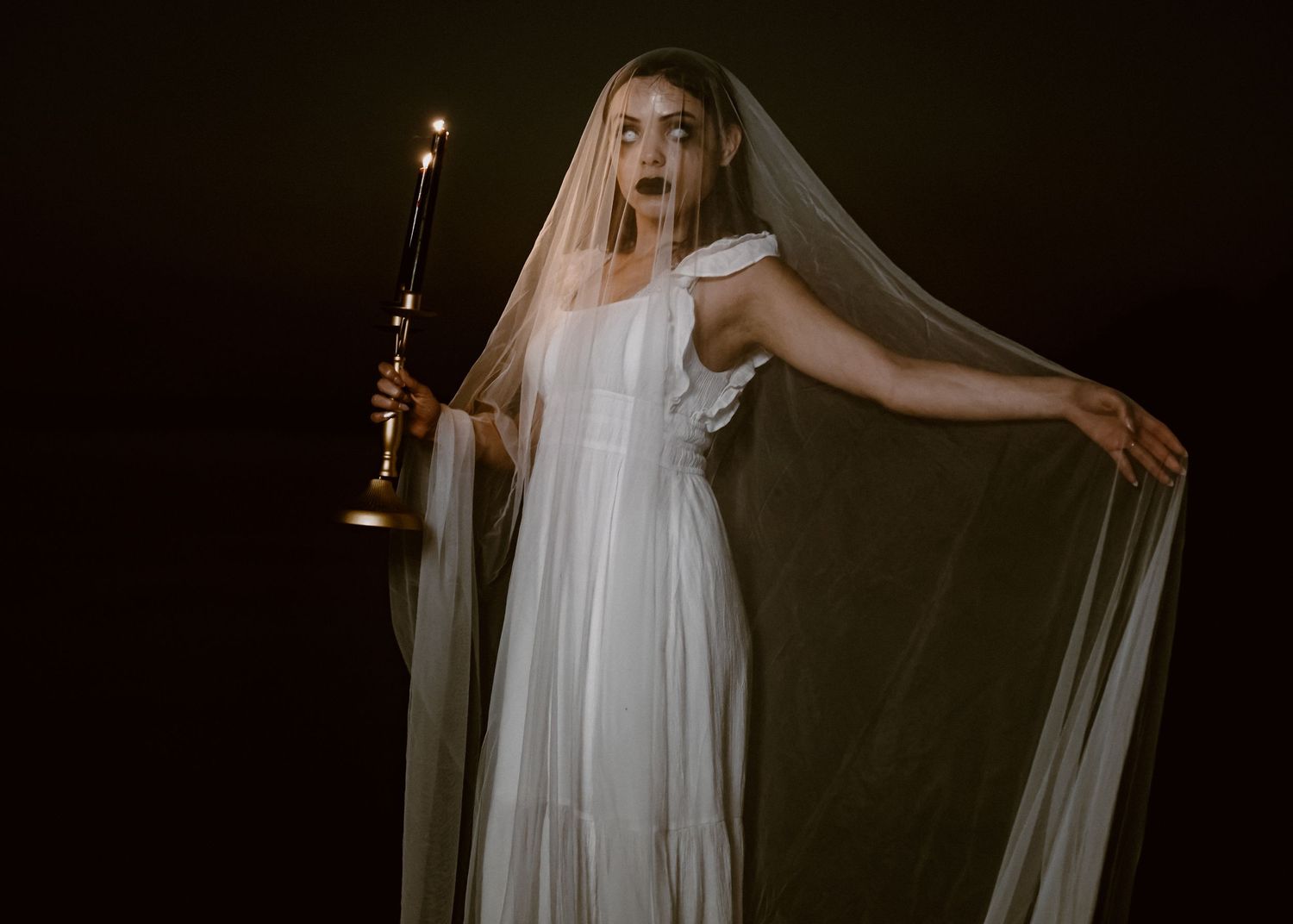Dramatic dark photoshoot featuring flowing white fabric and candlelight creating ethereal atmosphere.