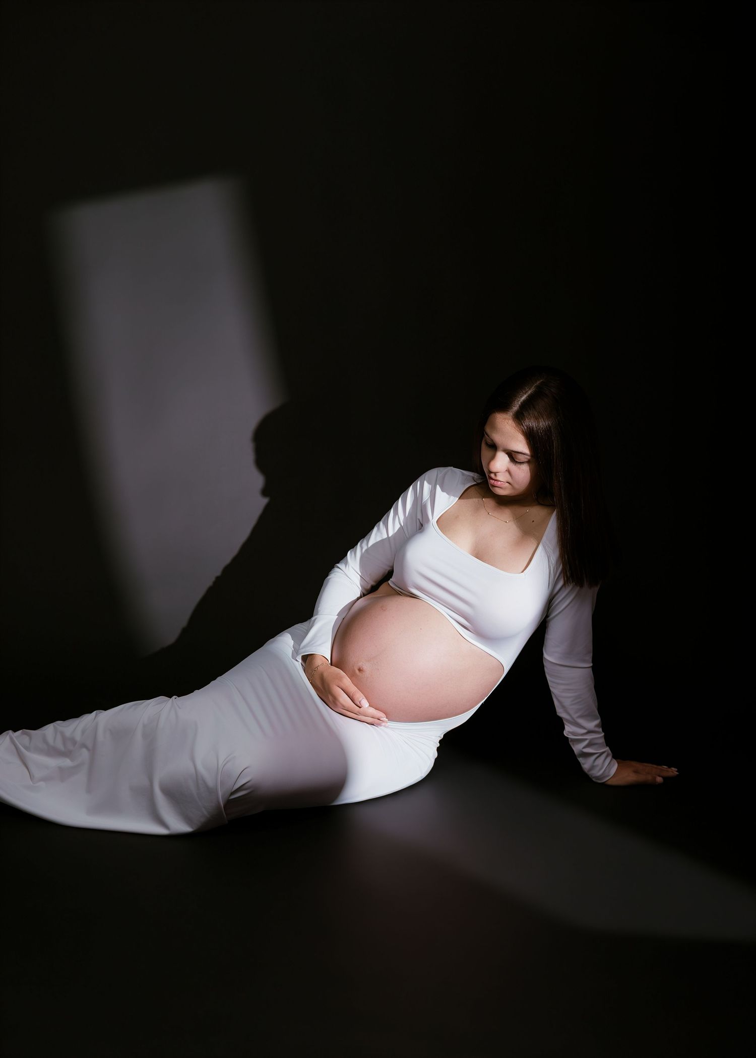 Pregnant woman in white dress sitting gracefully on floor against dark background, cradling her baby bump.