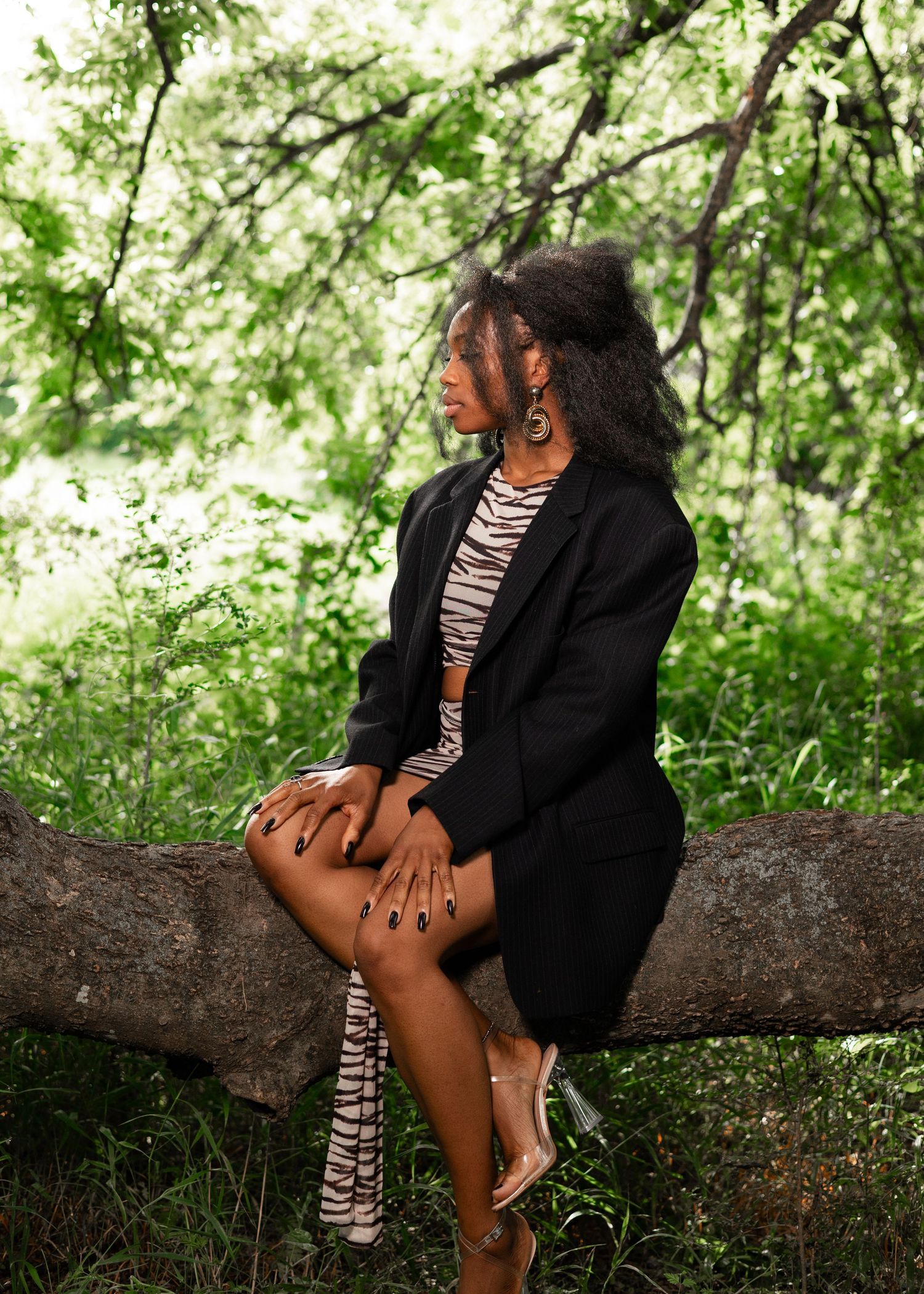 Woman in black blazer and zebra print outfit sits on a tree branch in a lush green forest setting.