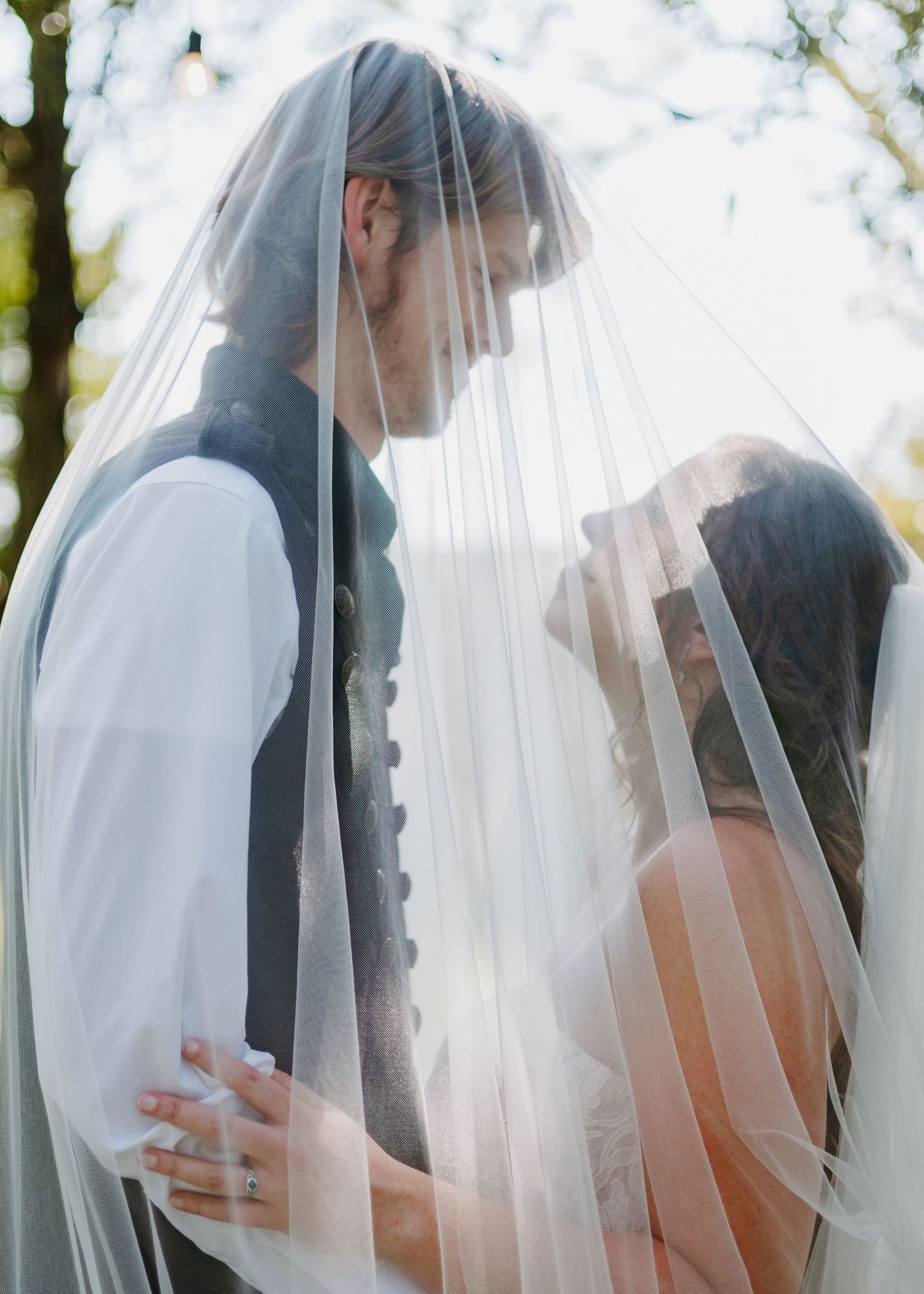 A bride and groom share an intimate moment under a flowing white wedding veil outdoors.