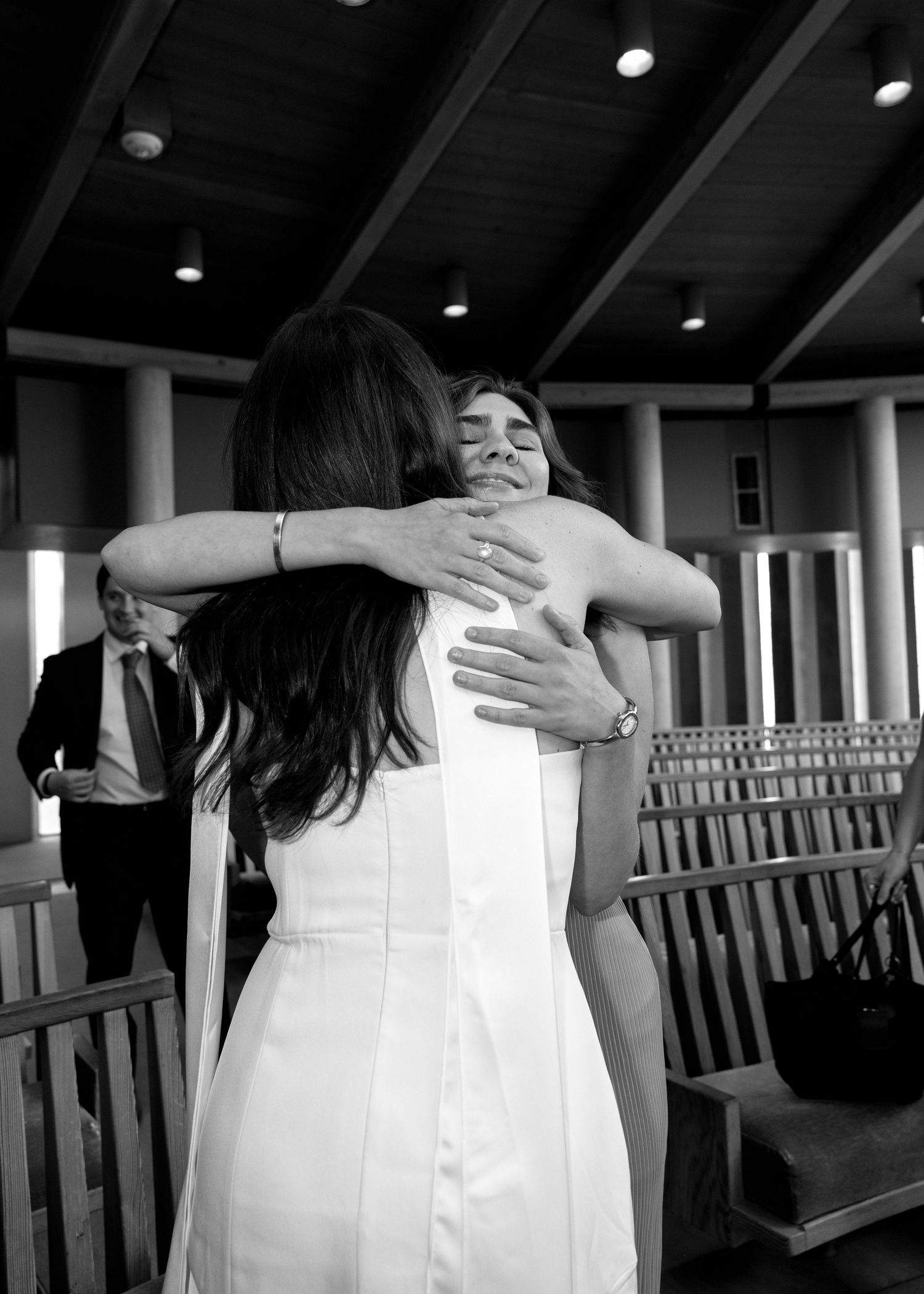 Two women share an emotional hug inside a church during a wedding ceremony in black and white.